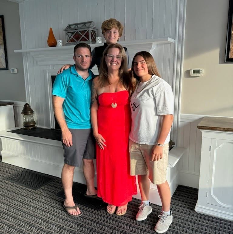 Four people standing indoors, a woman in a red dress in the center, two men and a girl around her, smiling.