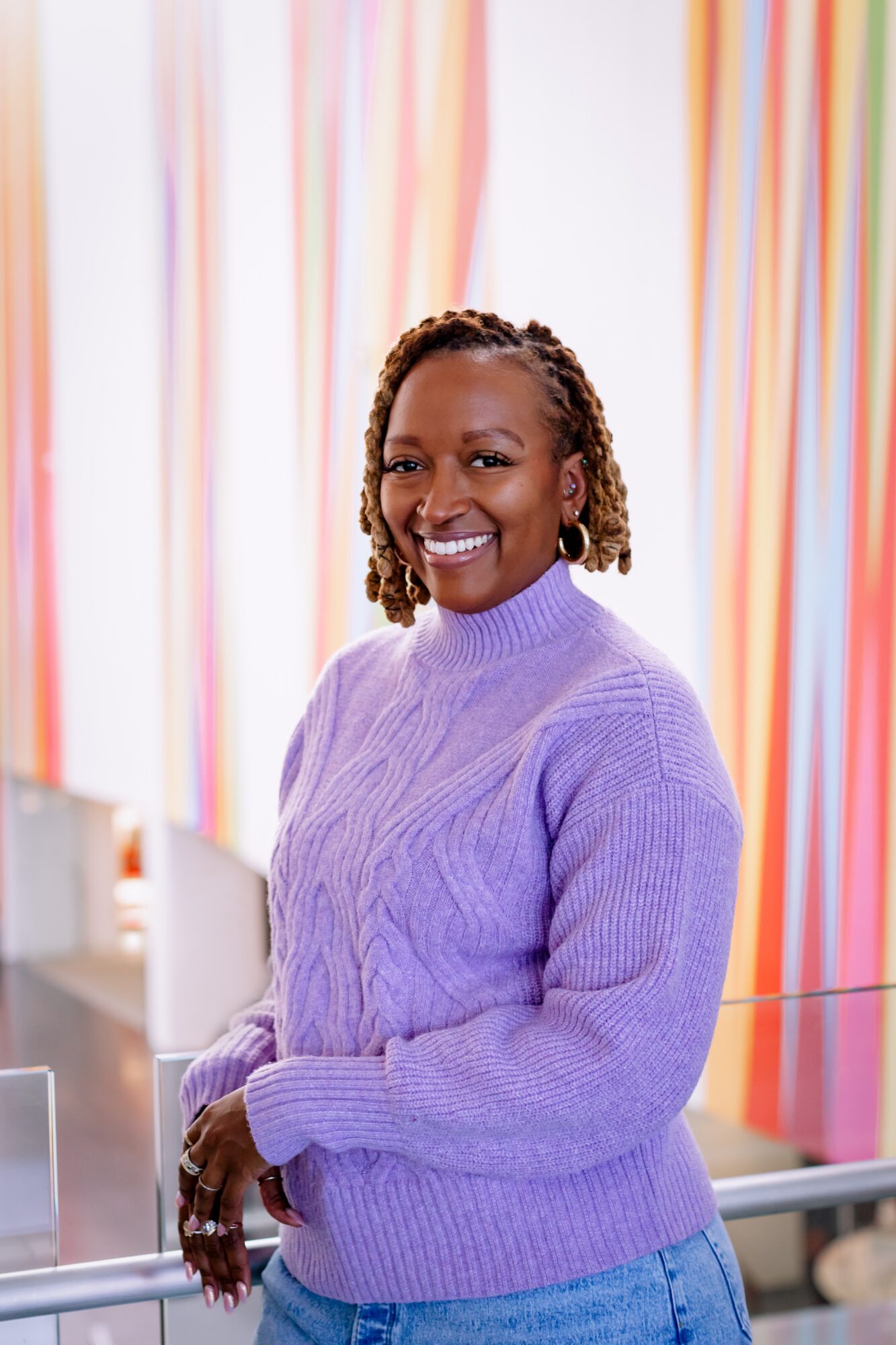 Smiling woman with curly hair wearing a purple sweater, standing indoors with colorful curtains in the background.