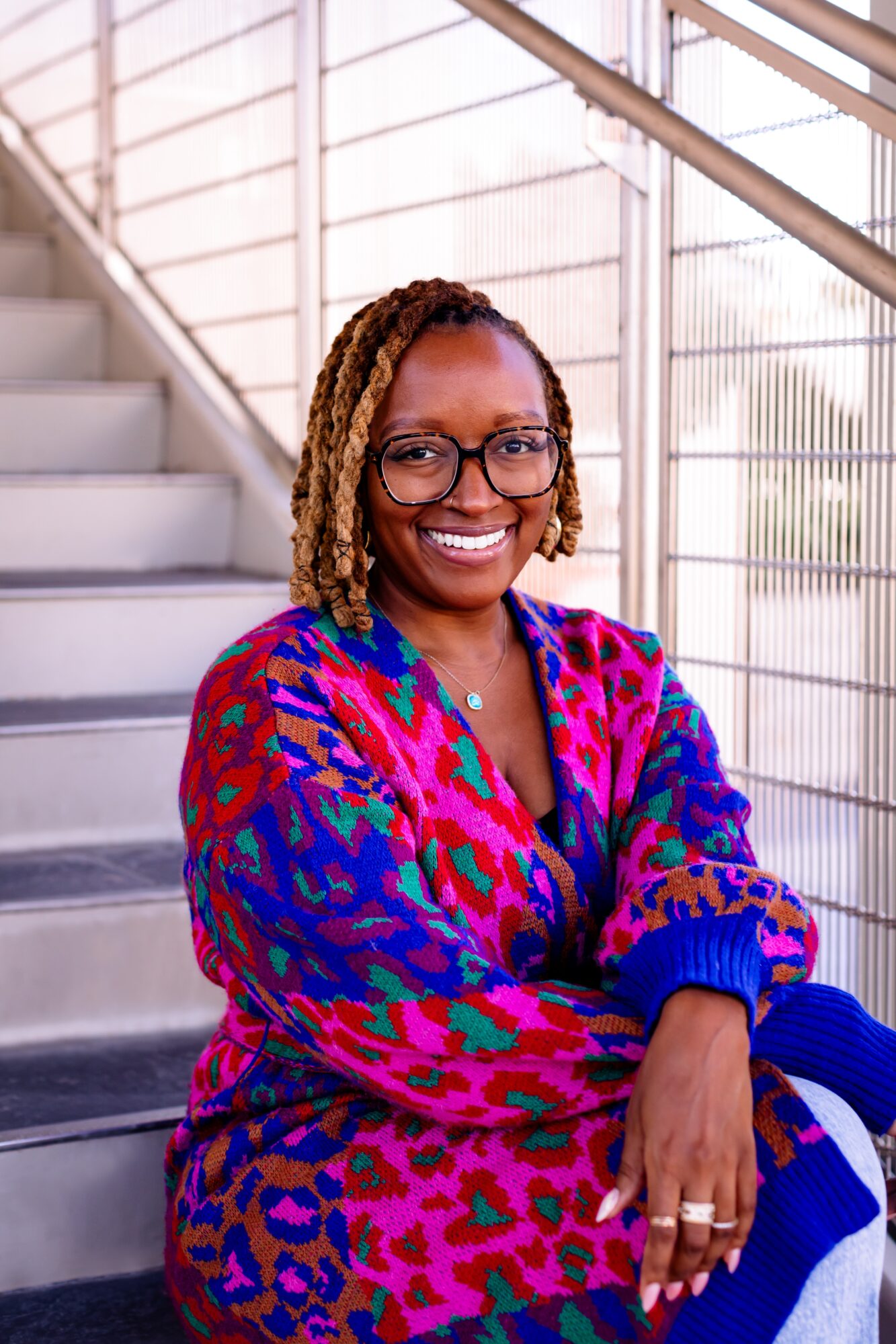 Smiling woman with glasses and dreadlocks sitting on stairs, wearing a colorful patterned sweater, in a modern building.