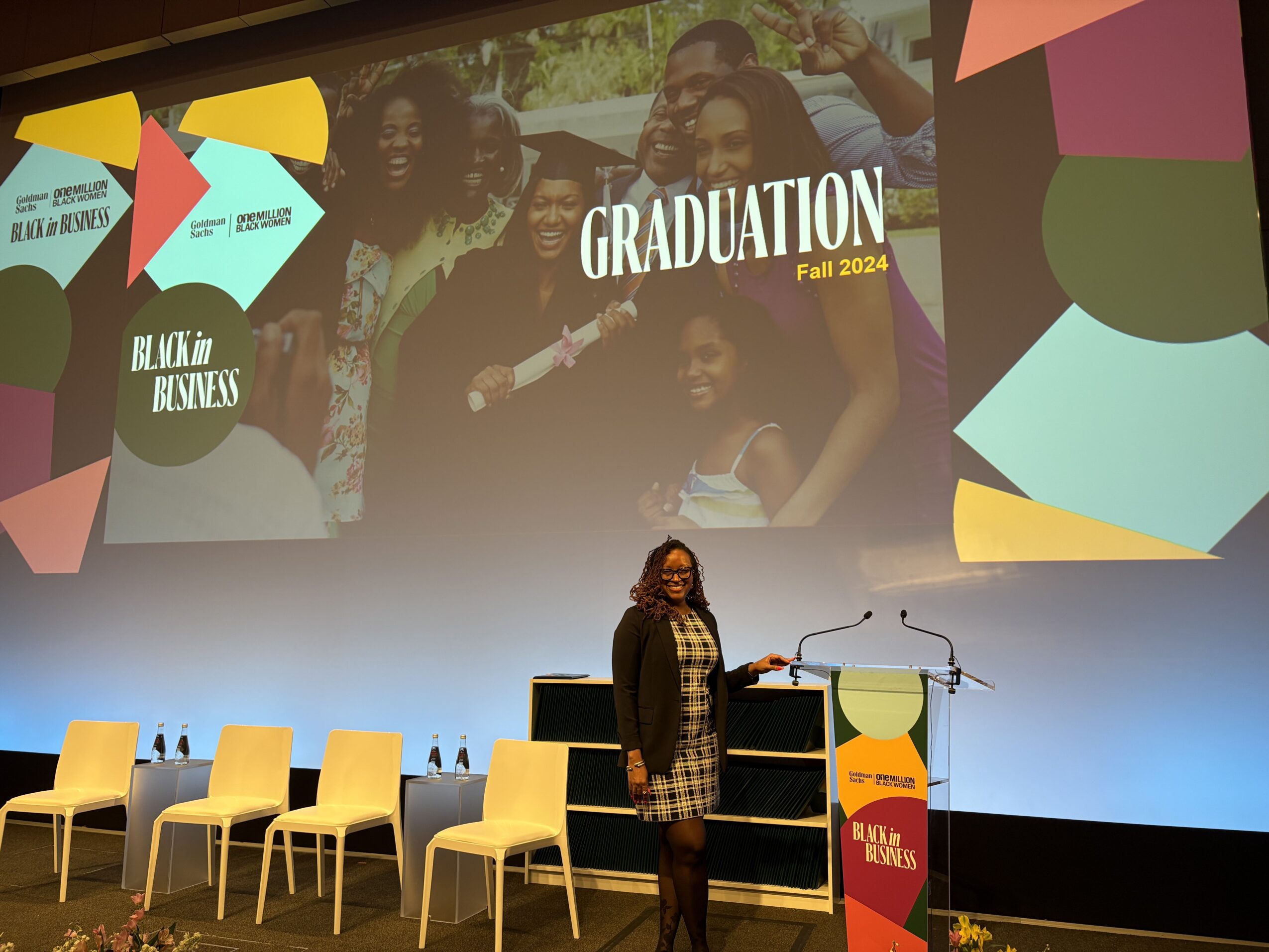 Woman standing on stage with a microphone, graduation event with chairs and a large screen displaying 'GRADUATION Fall 2024'.