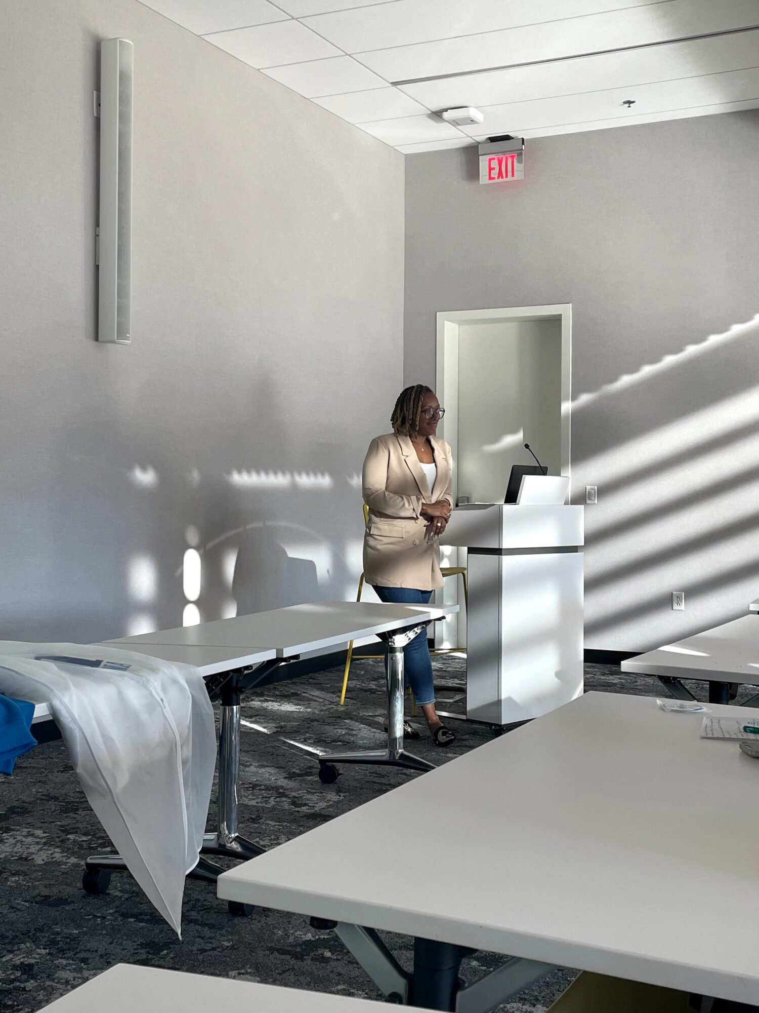 A woman stands at a white podium with a laptop in a room with gray walls and tables, sunlight creates striped shadows.