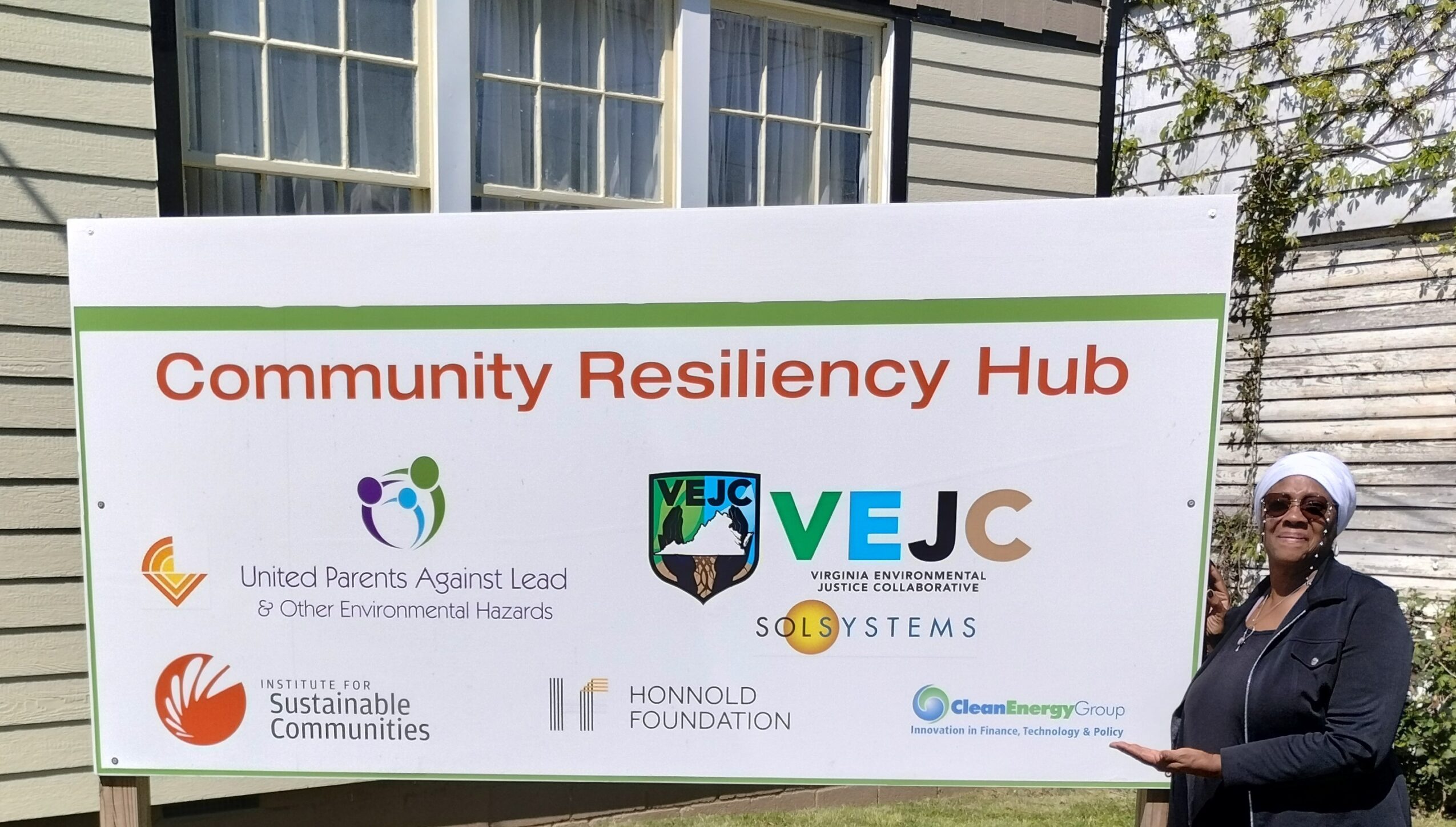Woman standing next to a large sign for Community Resiliency Hub with logos of organizations, outdoors, with buildings and trees in background.
