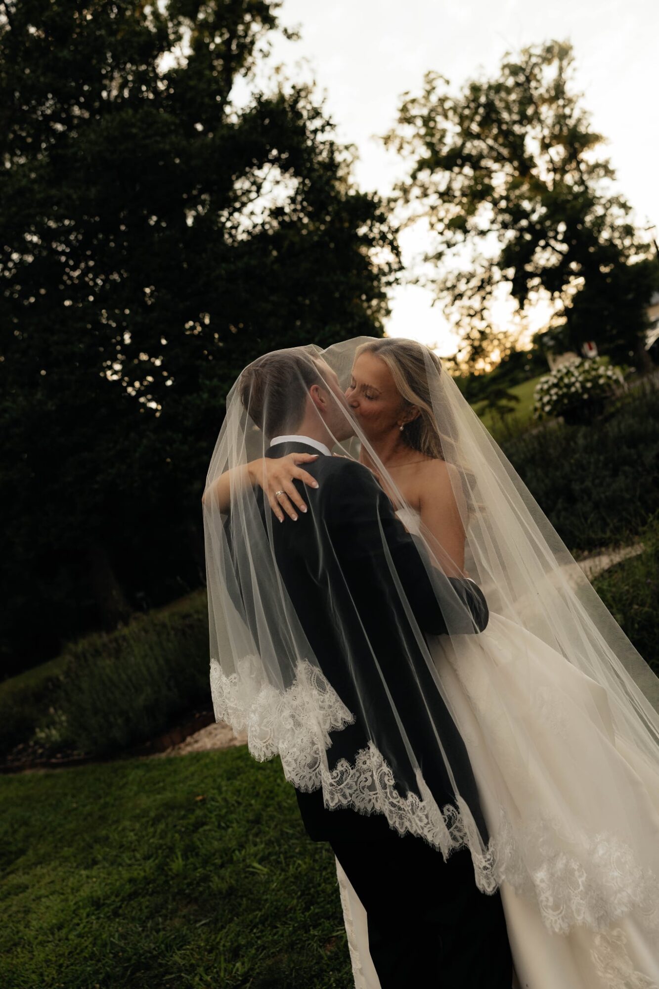 A bride and groom embrace outdoors, with trees and sky in the background, both smiling and holding each other.