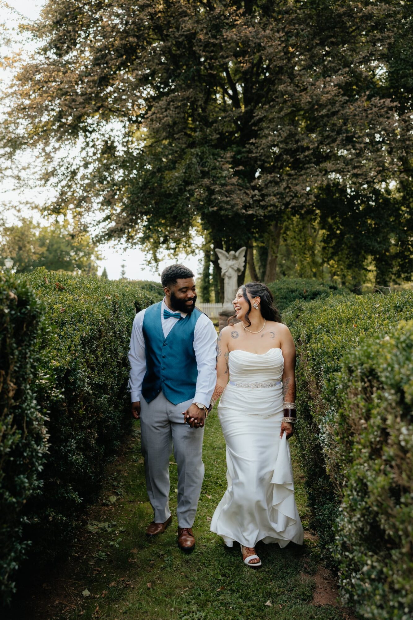 A couple holding hands walking outdoors on a grassy path with trees and bushes around them.