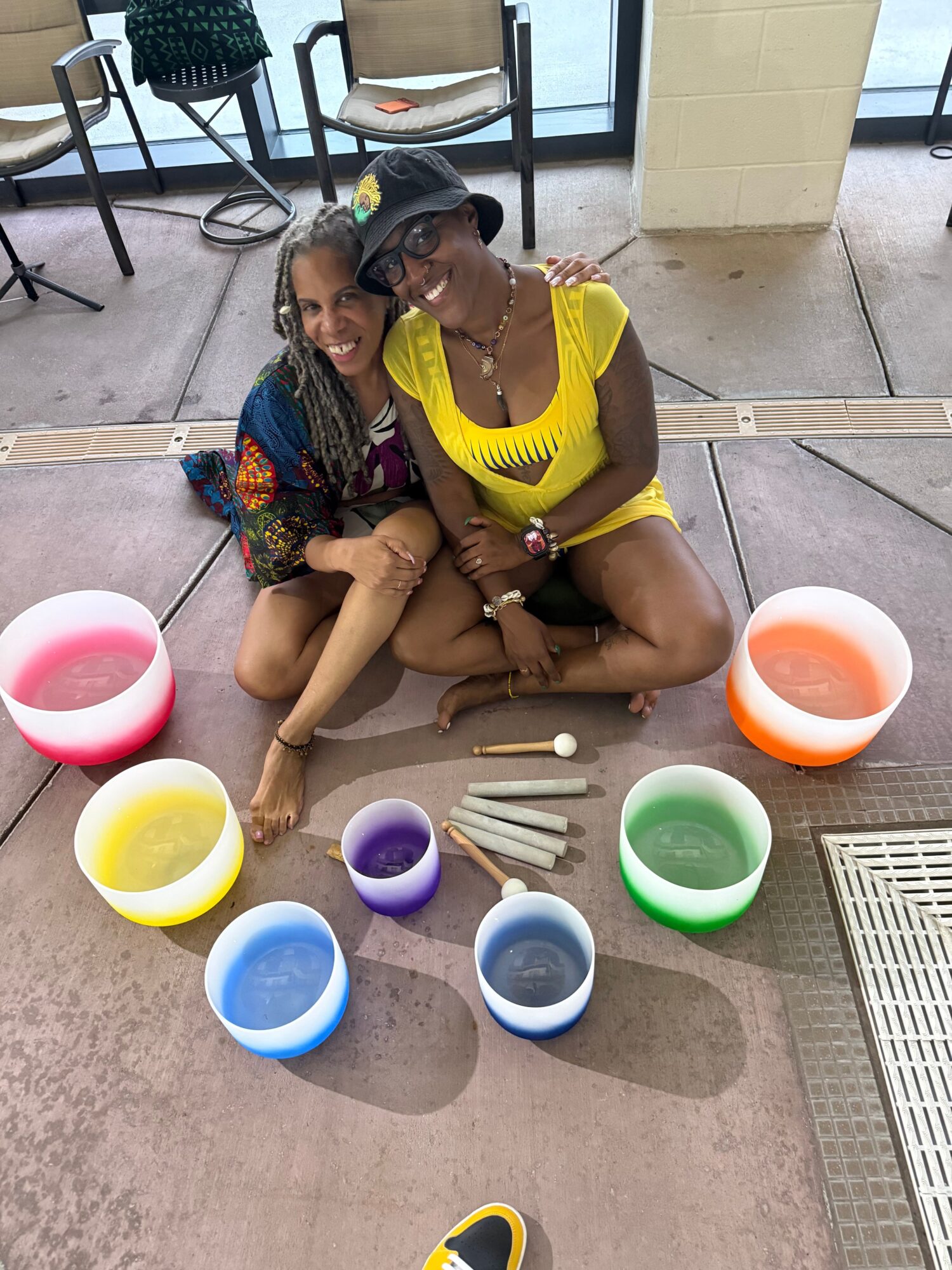 Two women sitting on the floor surrounded by colorful bowls and wooden sticks, engaging in an activity indoors.