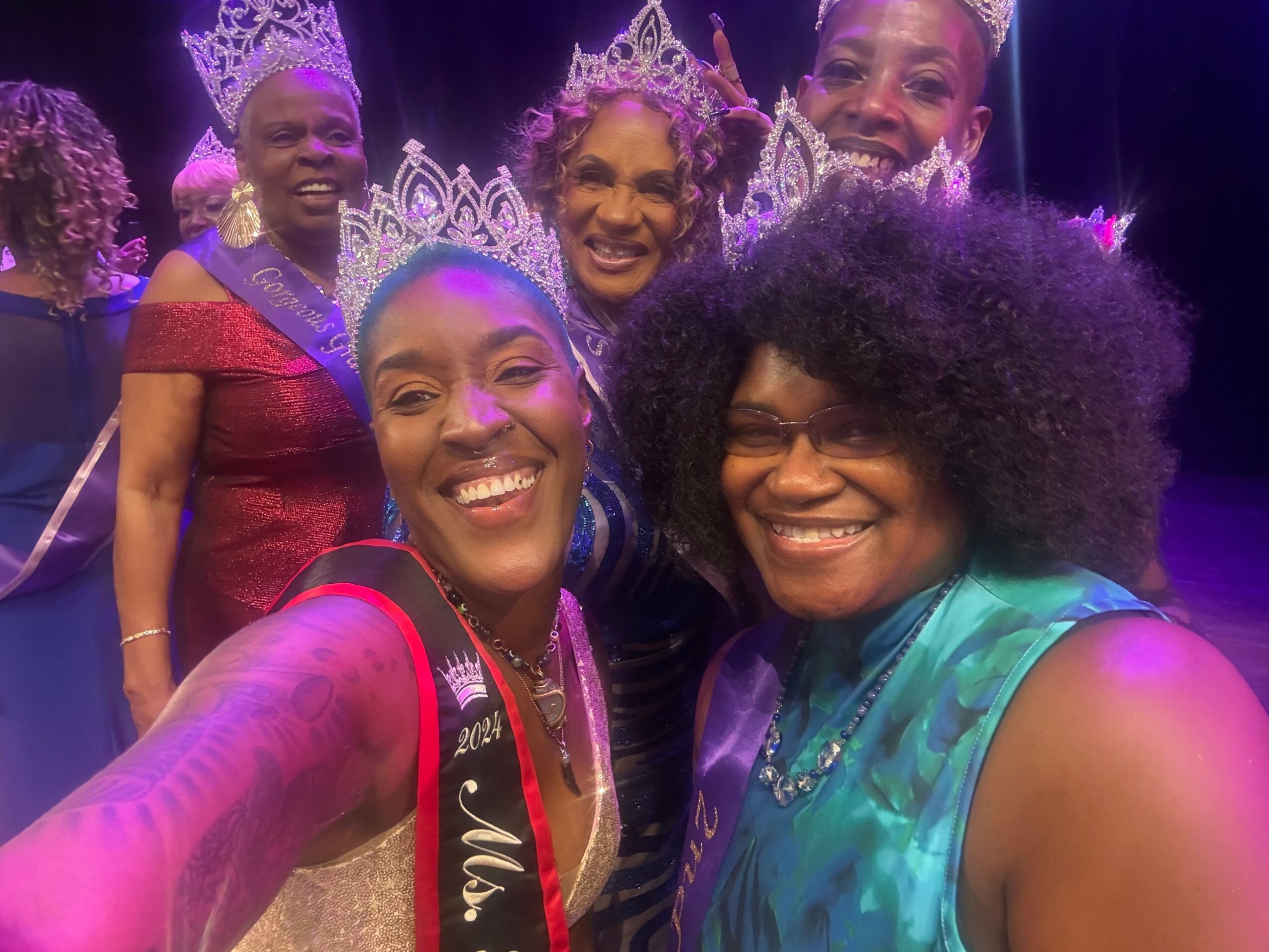 Four women in colorful dresses and tiaras smiling, celebrating at an event, with purple lighting in the background.