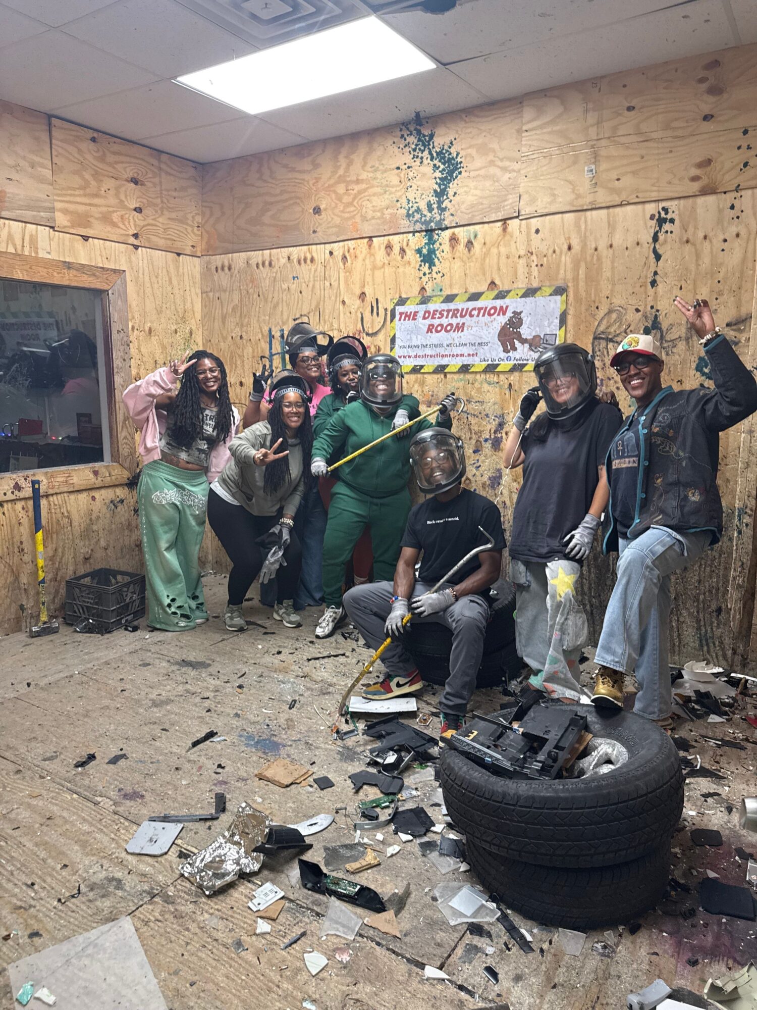 Group of people in a room with debris, some standing and some kneeling, with a tire and broken objects on the floor.
