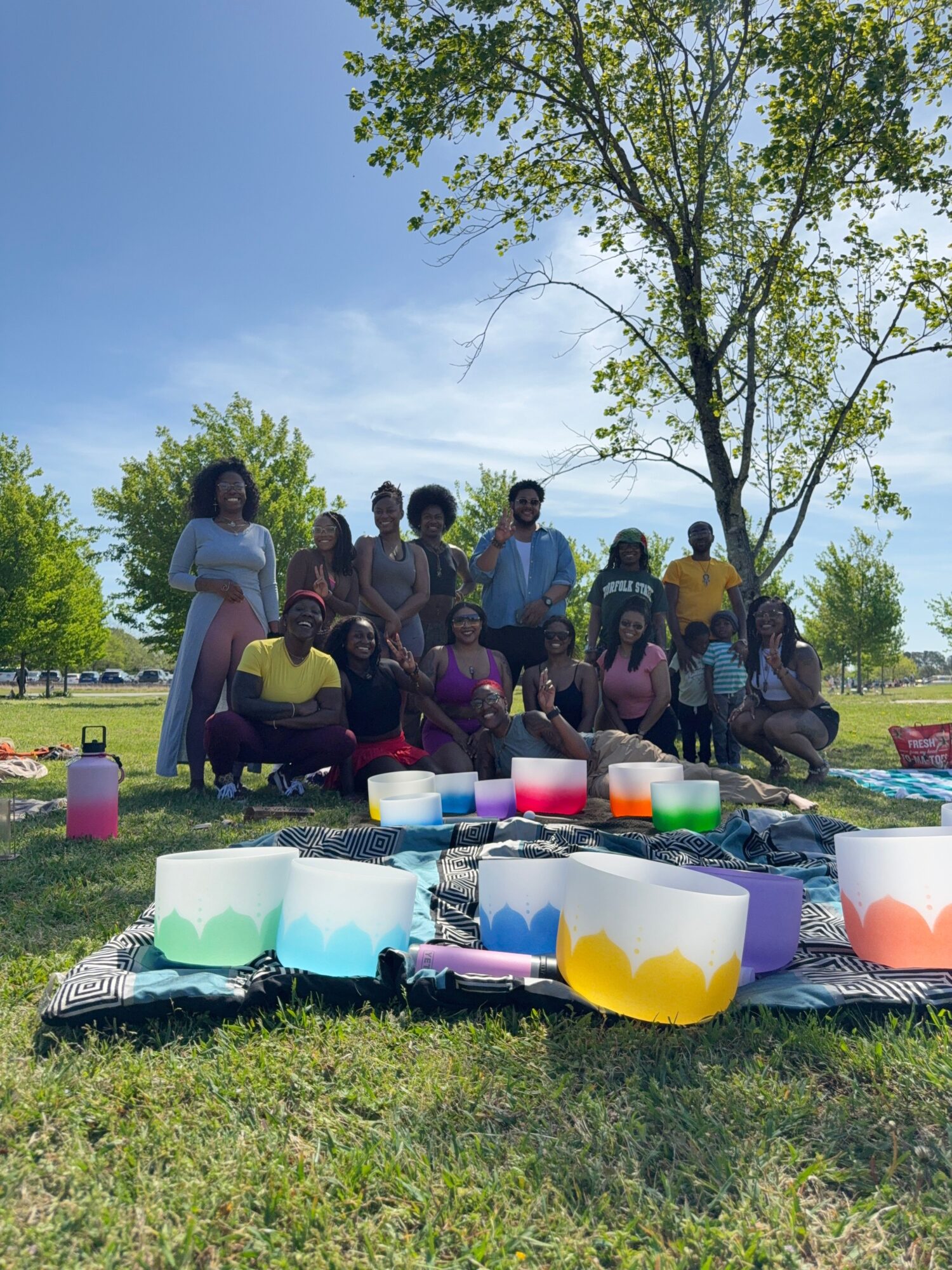 Group of people outdoors with a table of colorful food and drinks, trees and blue sky in background.
