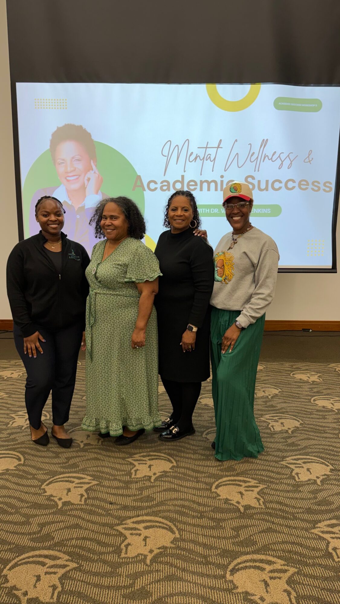 Four women standing together in front of a presentation screen, smiling, in a conference room.