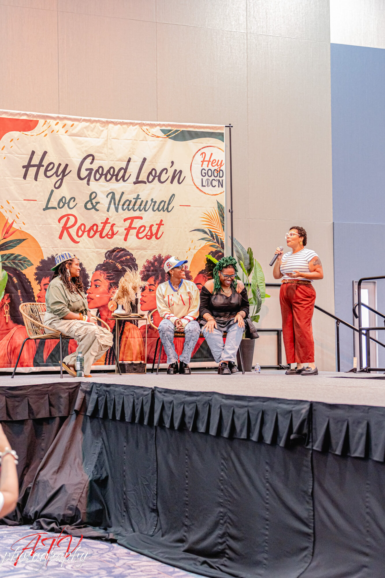 Six people seated on stage with a woman standing and speaking, a banner behind them reads 'Hey Good Loc'n Loc & Natural Roots Fest'.