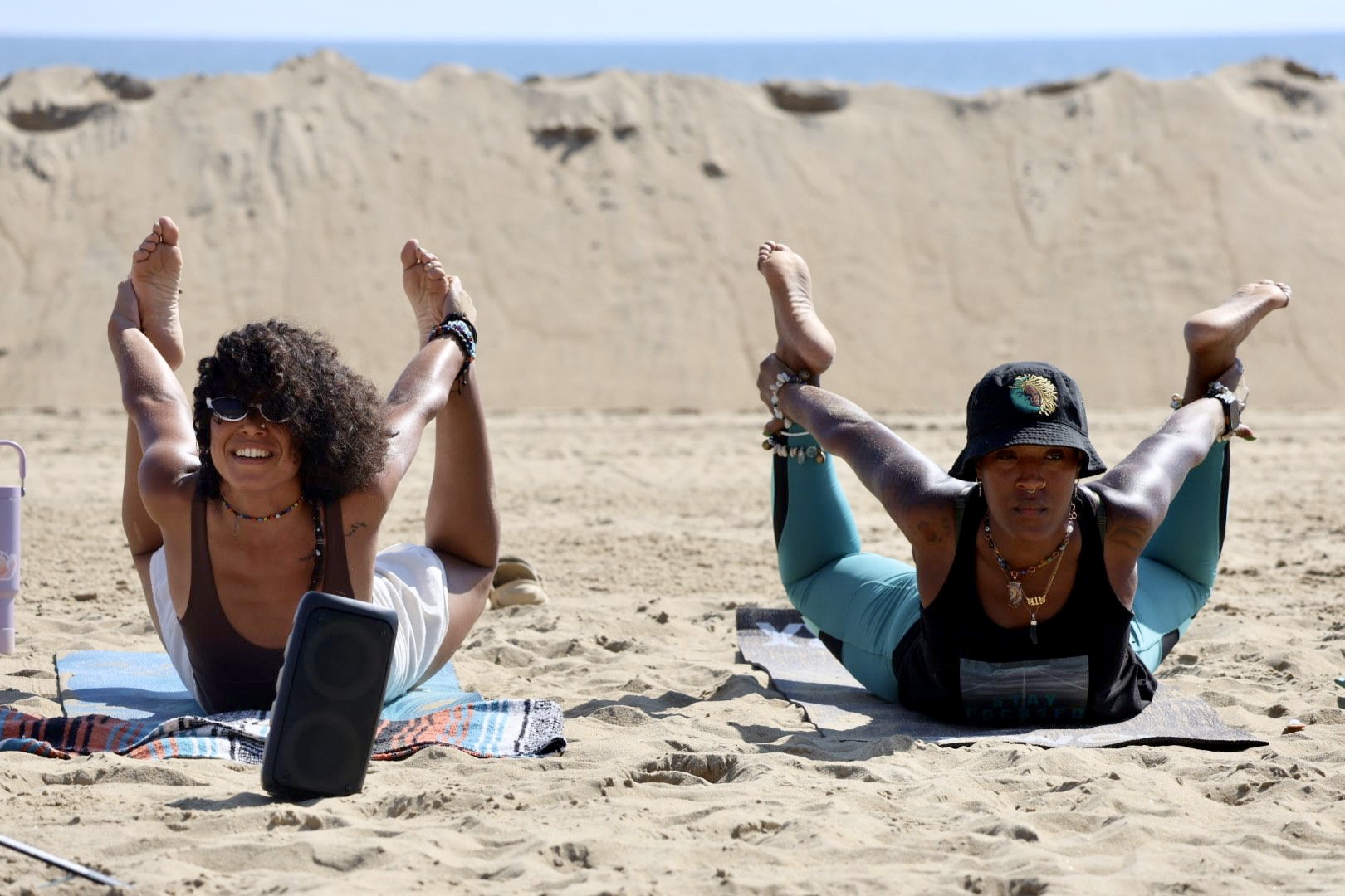 Two women practicing yoga on a sandy beach with a large sand dune in the background, both in a forward bend pose.