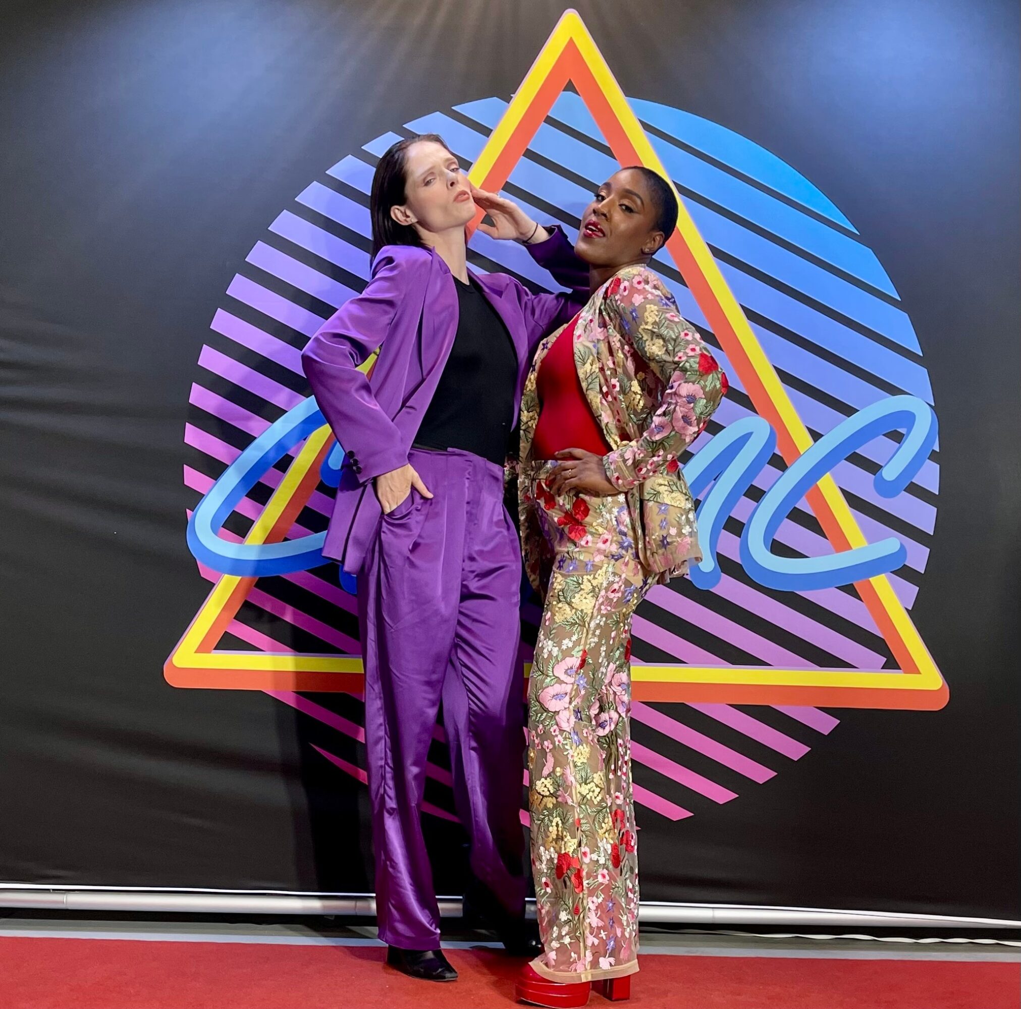 Two women stand in front of a colorful geometric background, one in purple and the other in floral attire, striking poses.