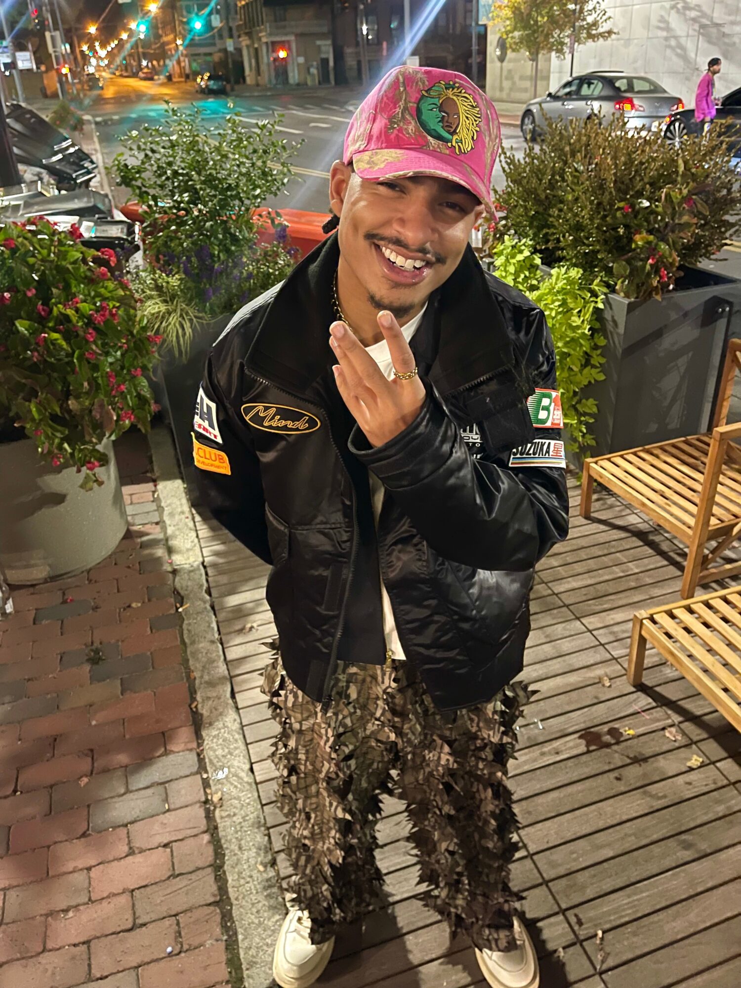 Person smiling, wearing a pink cap, black jacket, and patterned pants, standing outdoors at night with plants and benches nearby.
