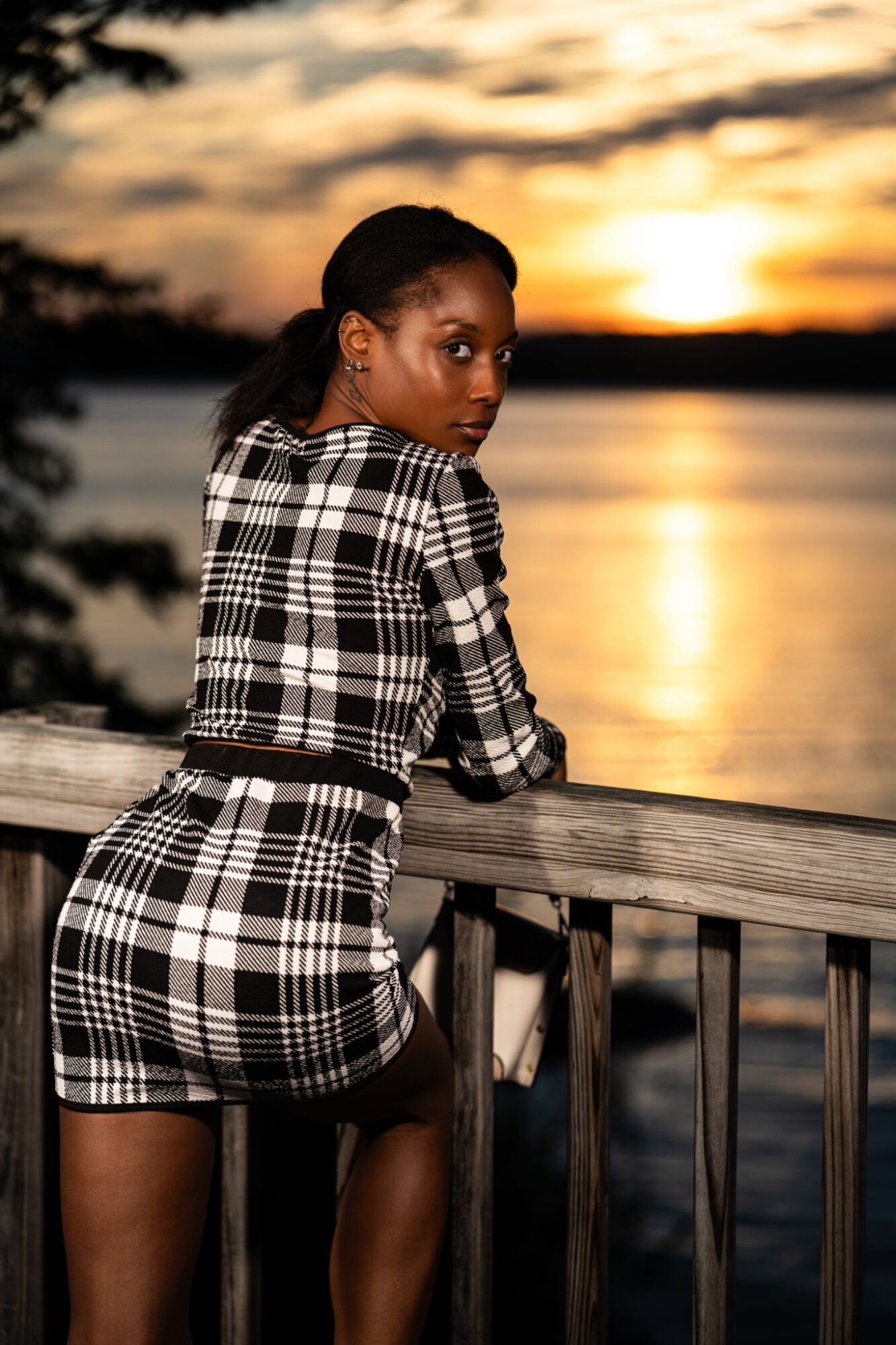 Woman in plaid outfit leaning on a railing by a lake during sunset, with trees and sky in the background.
