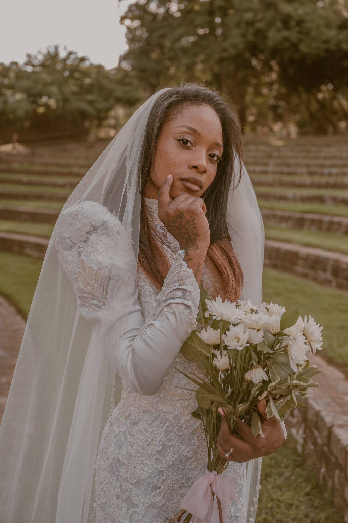 Woman in wedding dress holding a bouquet of white flowers outdoors, with trees and steps in the background.