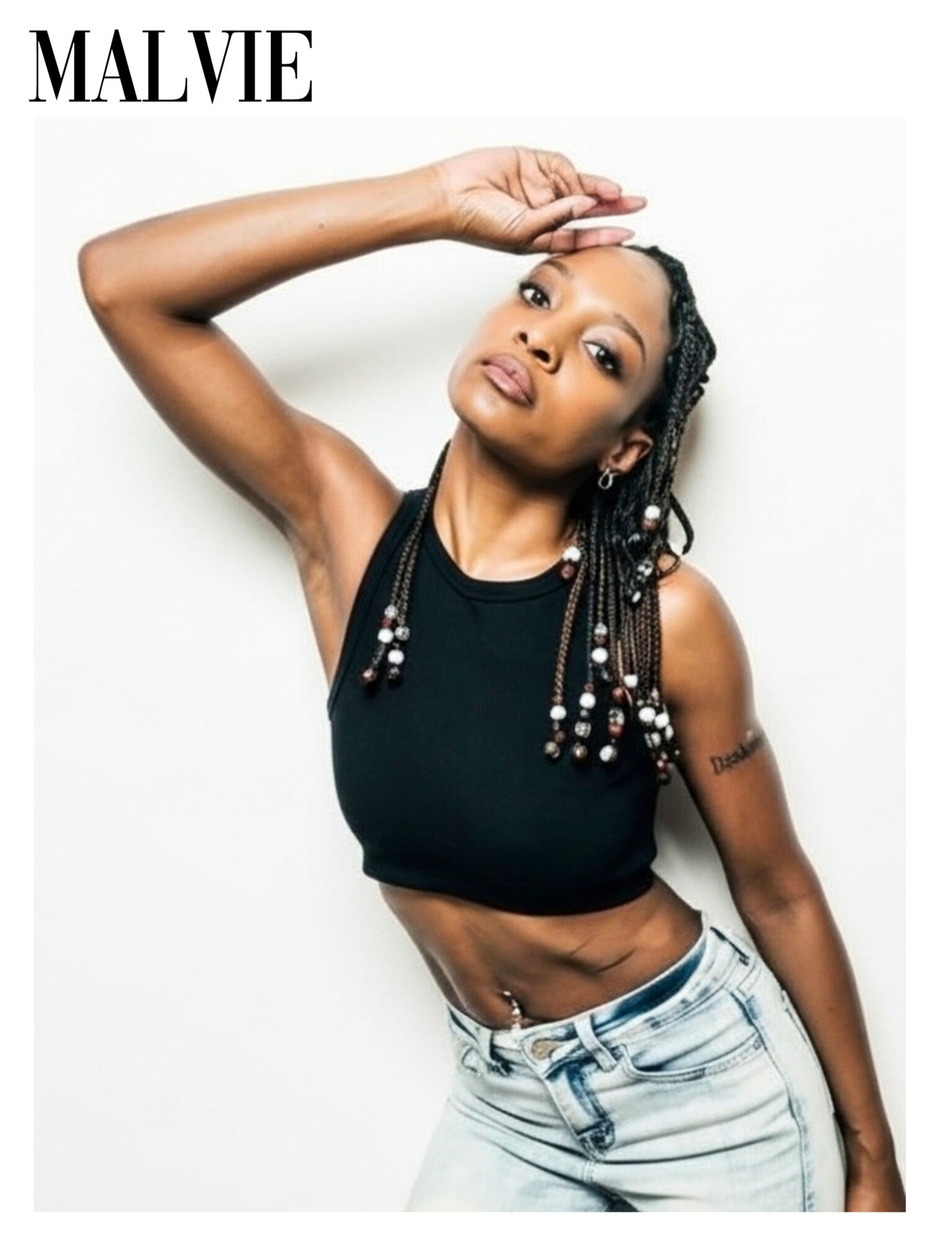Woman with braided hair wearing a black sleeveless top and light jeans, posing with her hand on her head.