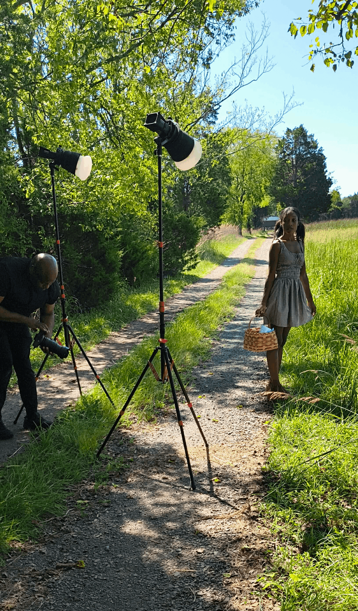 A girl in a dress holding a basket stands on a dirt path surrounded by greenery, with a photography setup nearby.