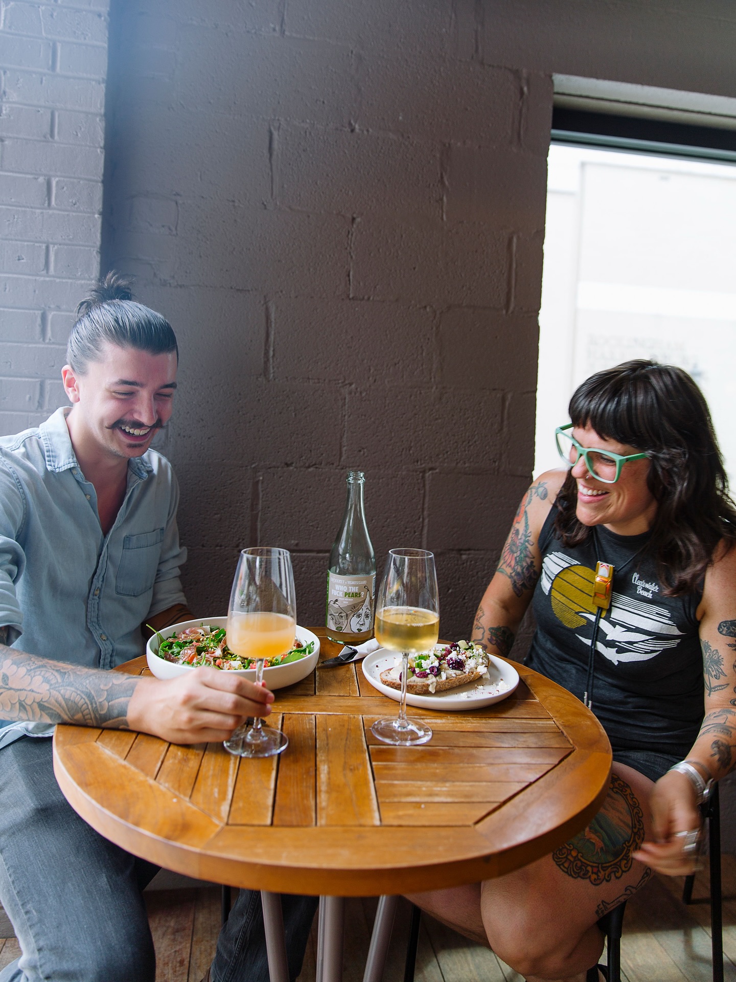 Two women sit at a round wooden table with drinks and food, smiling and talking indoors near a window.
