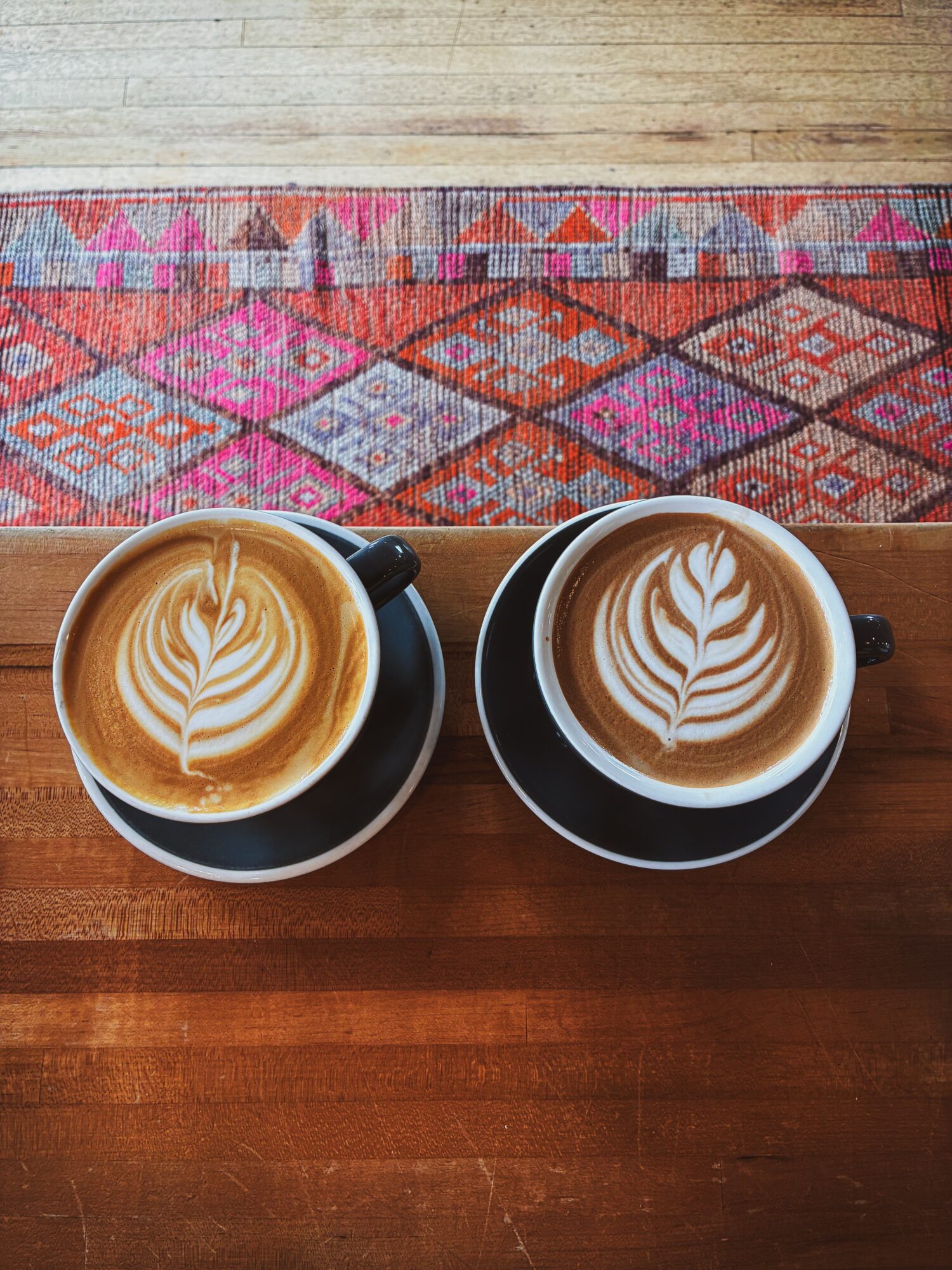 Two cups of coffee with latte art on a wooden table, with a colorful rug in the background.