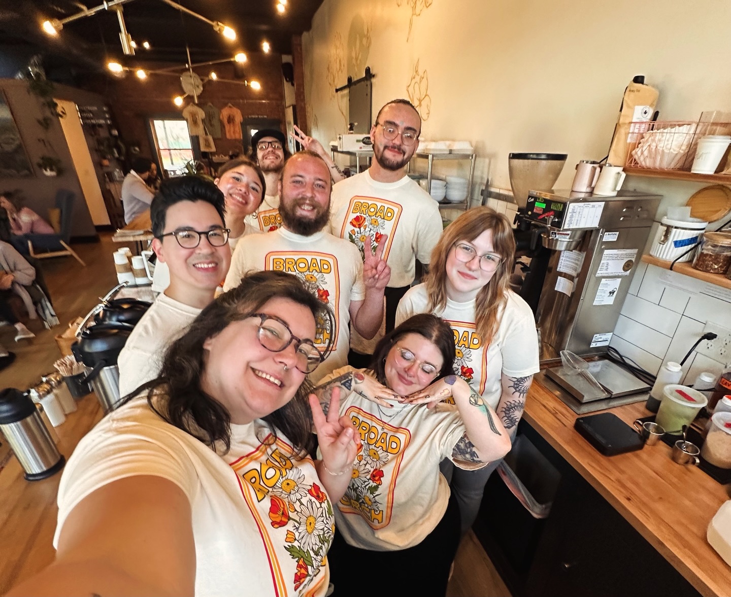 Group of seven people smiling and posing inside a restaurant, some making peace signs, with a counter and kitchen in the background.