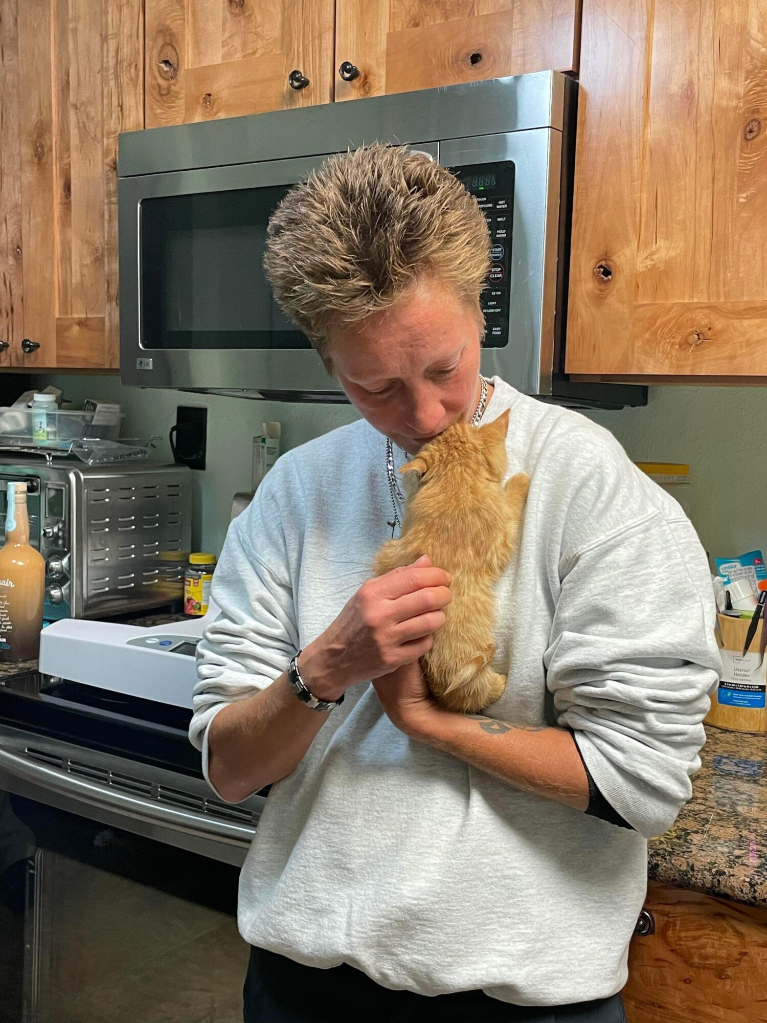 Person holding an orange kitten in a kitchen with wooden cabinets and a microwave above the stove.