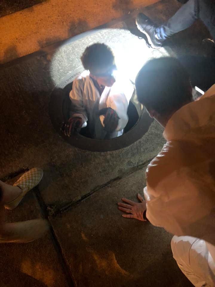 Person looking into a manhole with others nearby, illuminated by a bright light, on a dark street.