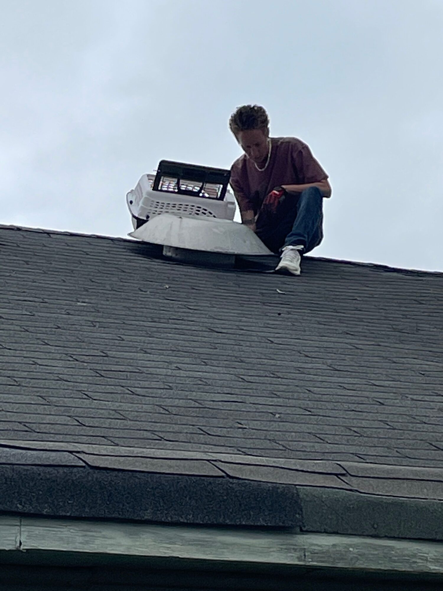 Person crouching on a rooftop with a white object, with a car and cloudy sky in the background.