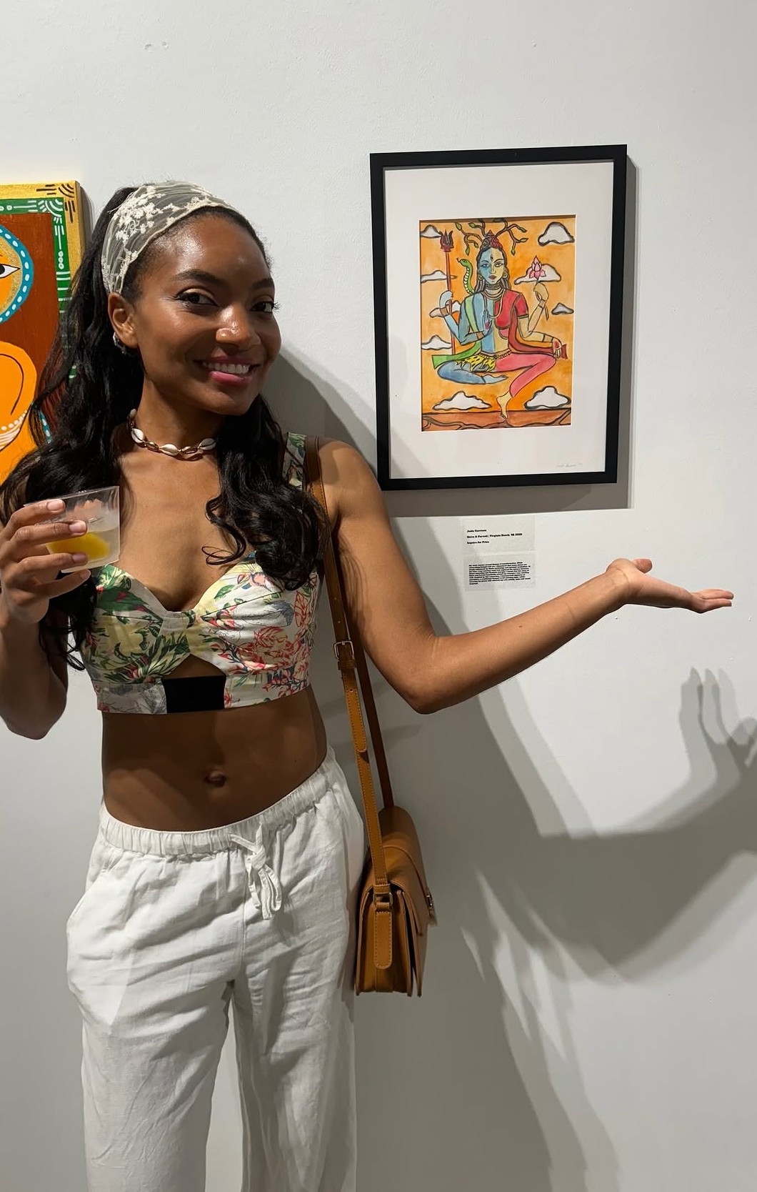 Young woman with long dark hair, wearing a floral crop top and white pants, standing in front of artwork, holding a drink.