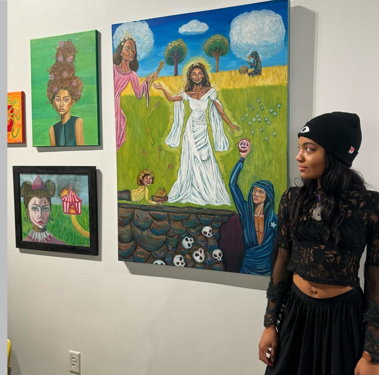 Young woman in black beanie and lace top stands in front of colorful paintings on gallery wall.