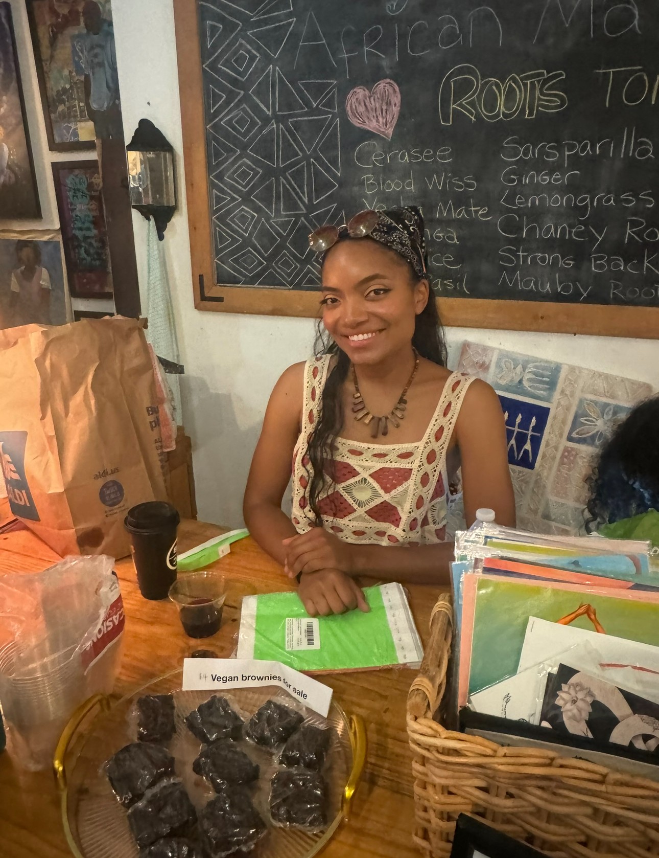 Smiling woman with braided hair and patterned dress sitting at a table with books and a basket, in front of a chalkboard.