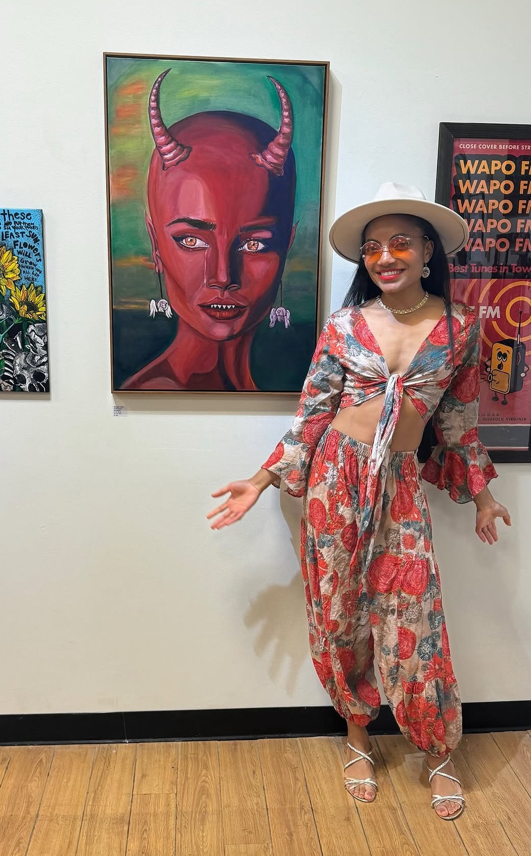 Woman in a floral dress and hat standing in front of colorful artwork on a white wall, smiling with arms outstretched.