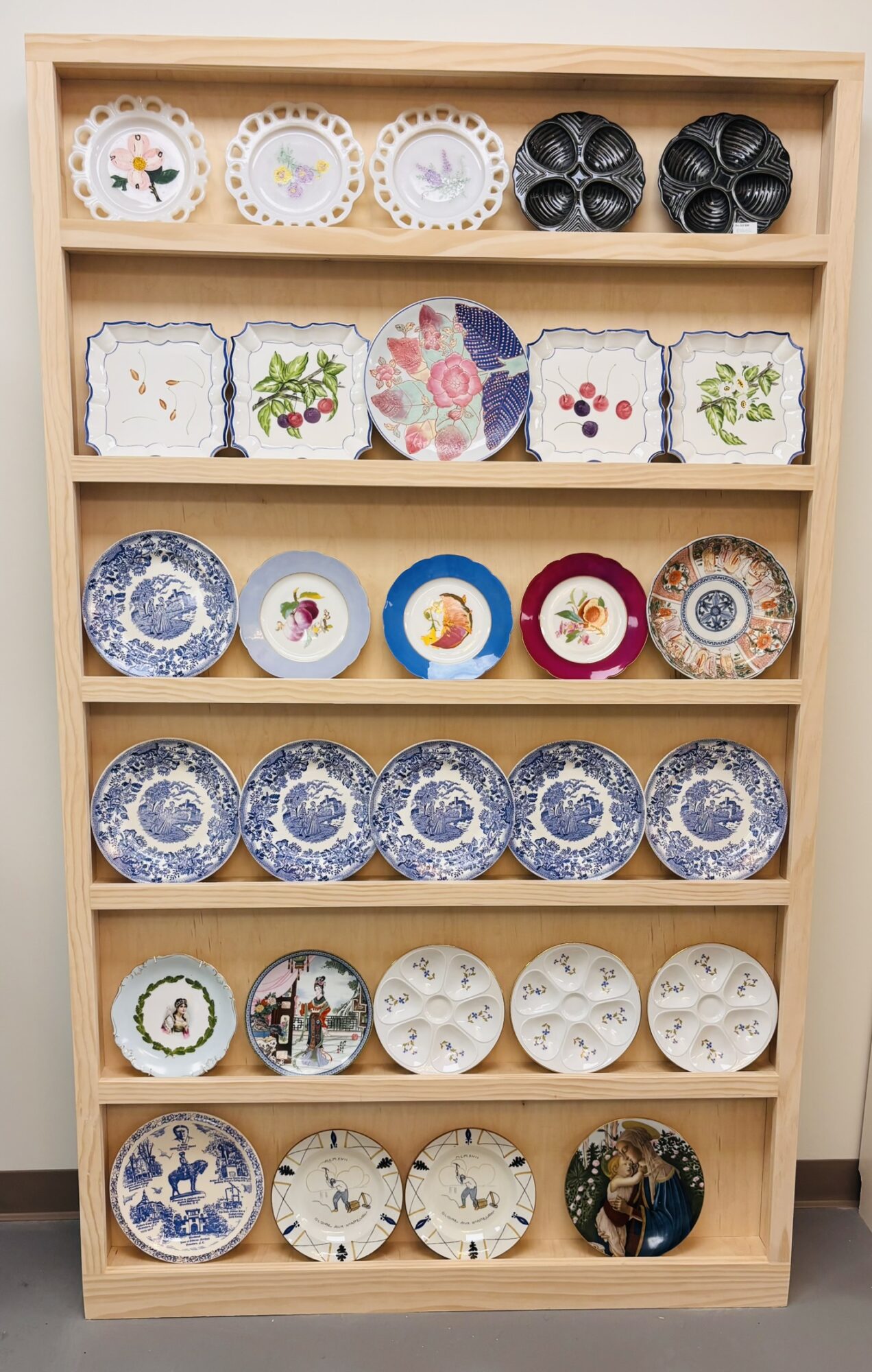 Wooden shelf with decorated plates and bowls arranged in rows, featuring floral, holiday, and abstract designs.