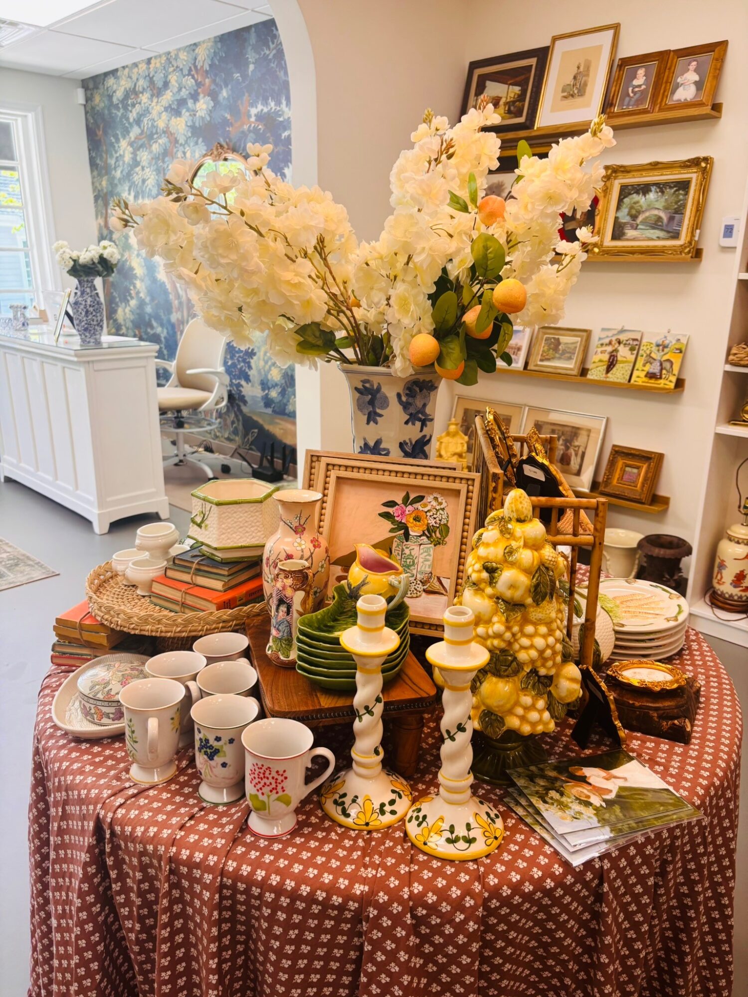 Room with a table filled with decorative items, cups, and flowers, and a wall with framed pictures, a window, and a blue wall.