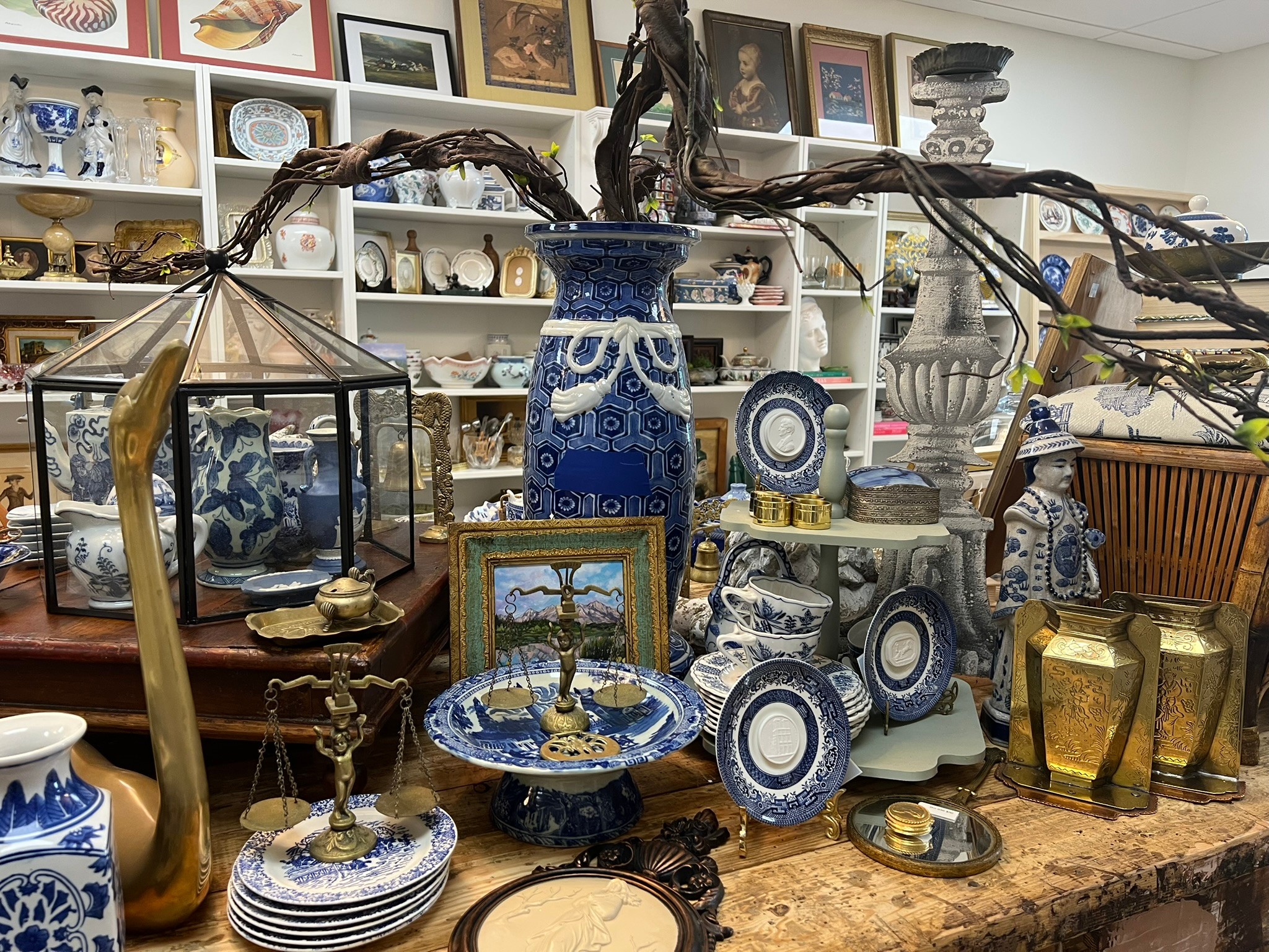 Display of blue and white ceramic plates, vases, and decorative items on a table with shelves in background.
