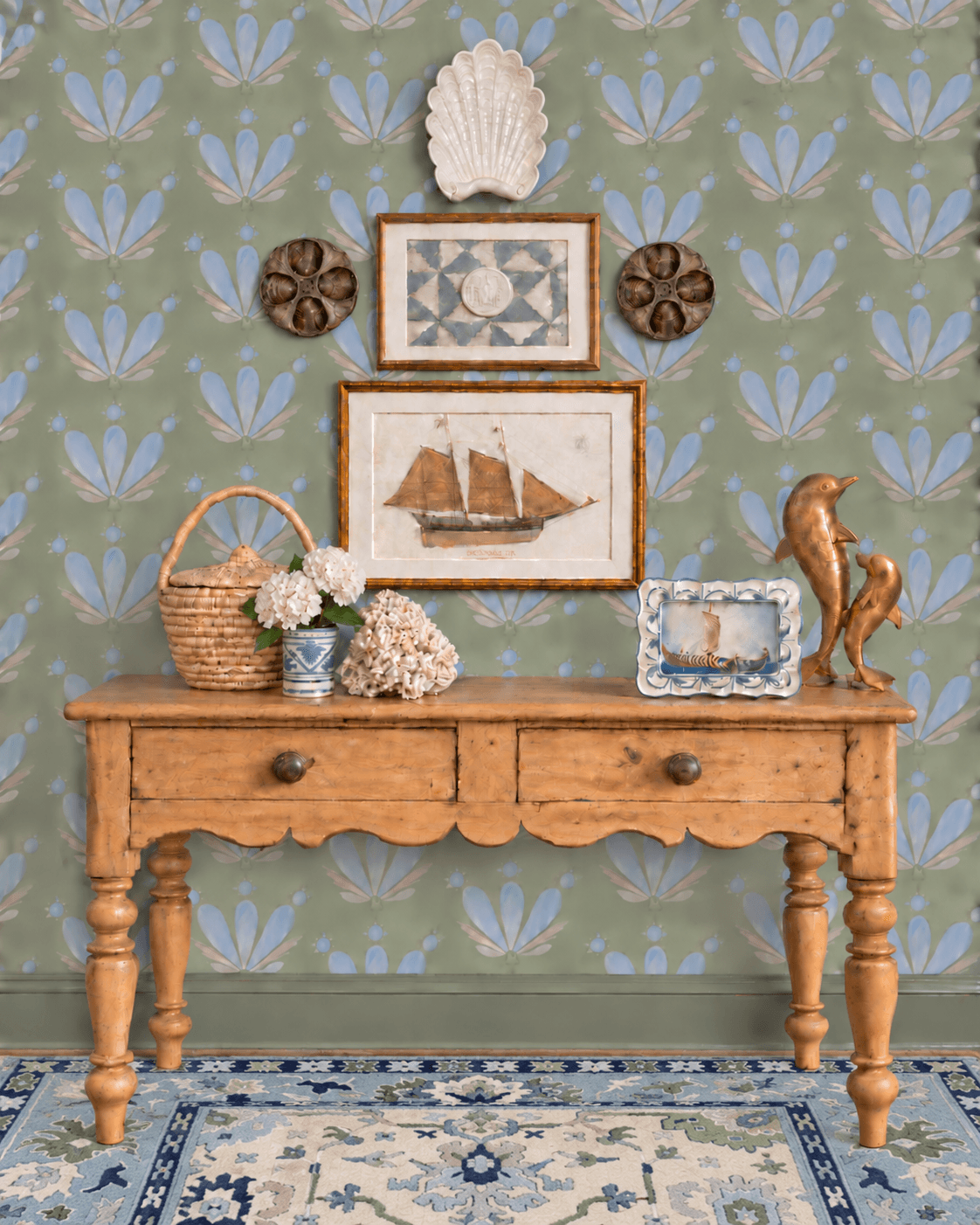 Wooden table with decorative items, framed pictures, basket, flowers, and a sculpture against a patterned wall and rug.