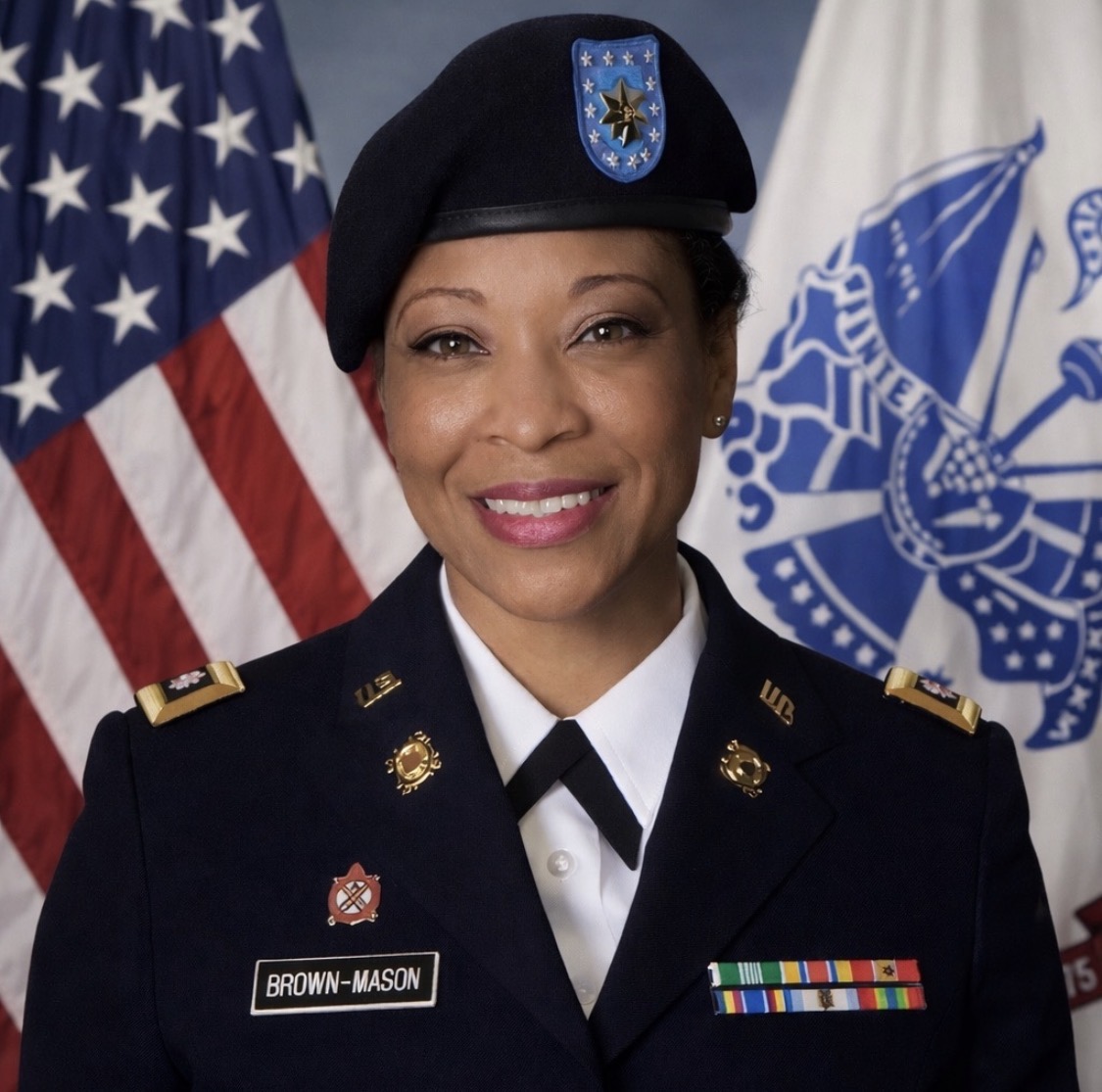 Woman in military uniform with flags in background, smiling, wearing a beret and medals.