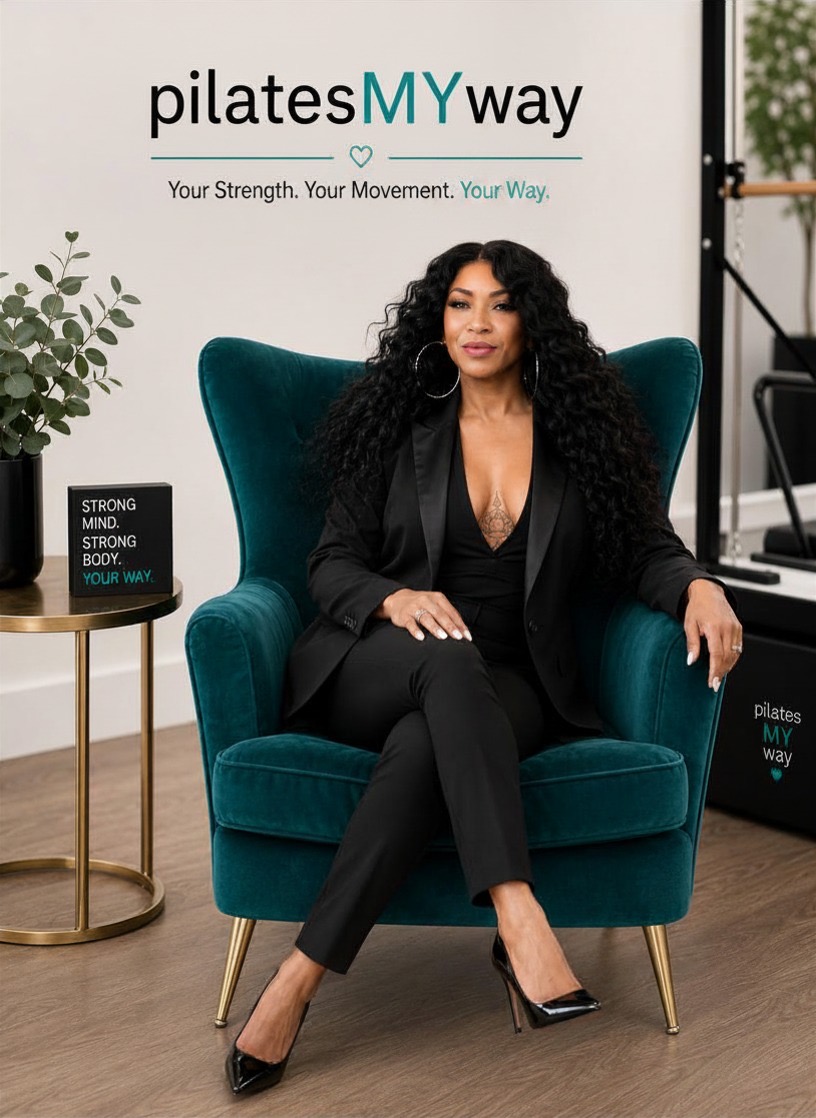 Woman with curly hair sitting in a teal armchair, wearing a black suit, in a room with a sign and plant.