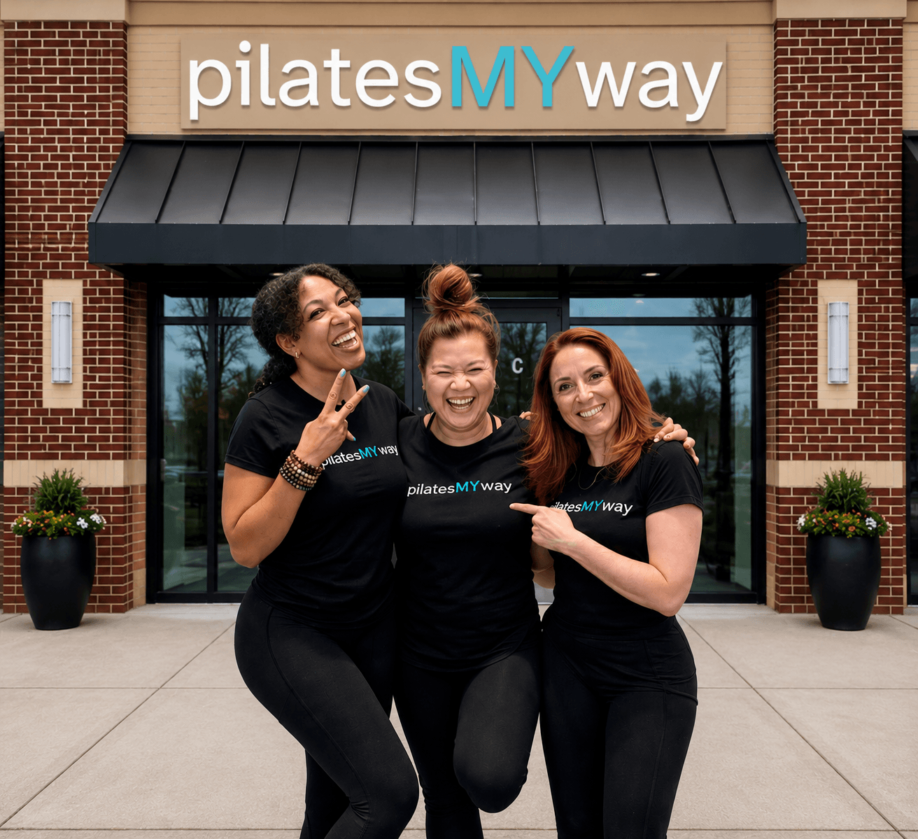 Three women in black shirts smiling and posing outside a Pilates studio with a sign that reads 'pilates MY way'.