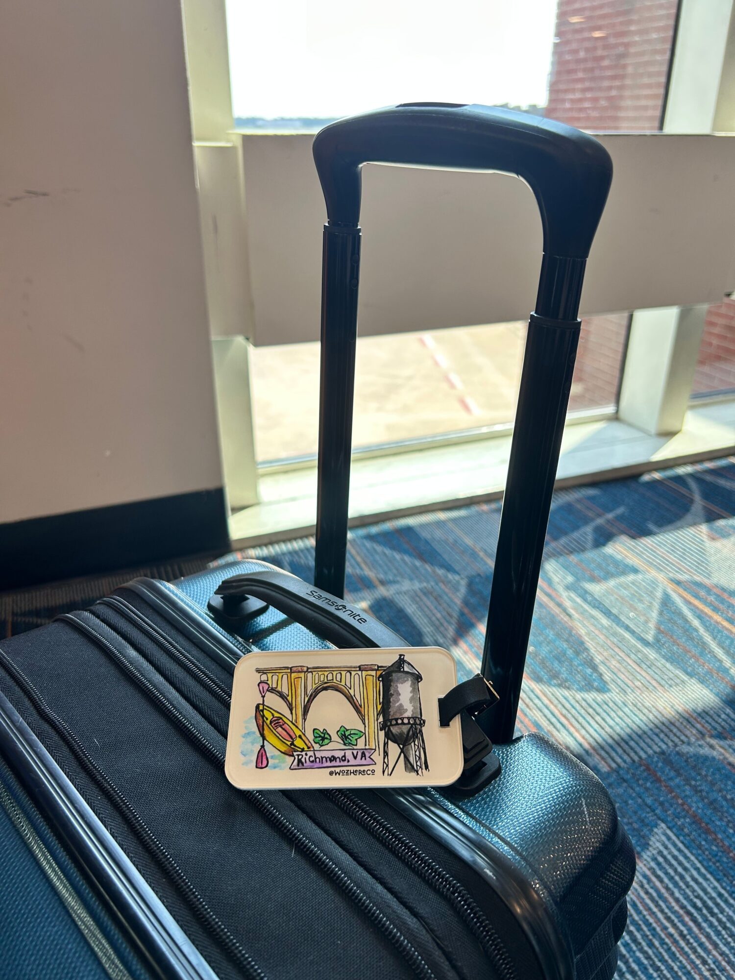 Black rolling suitcase with extended handle and a luggage tag, placed near a window with patterned carpet visible.