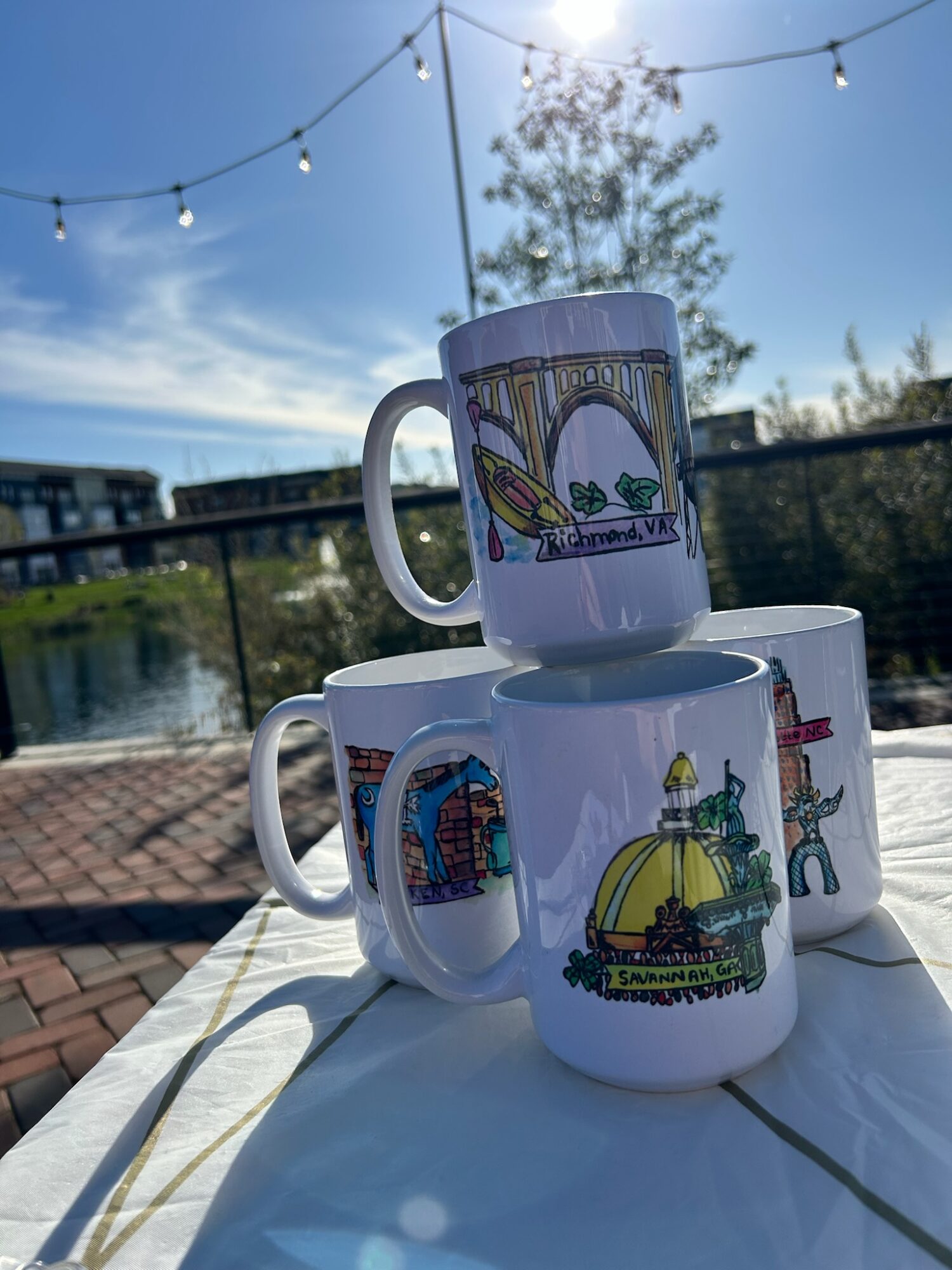 Three ceramic mugs with colorful illustrations stacked outdoors on a table near a body of water, with a string of lights overhead.