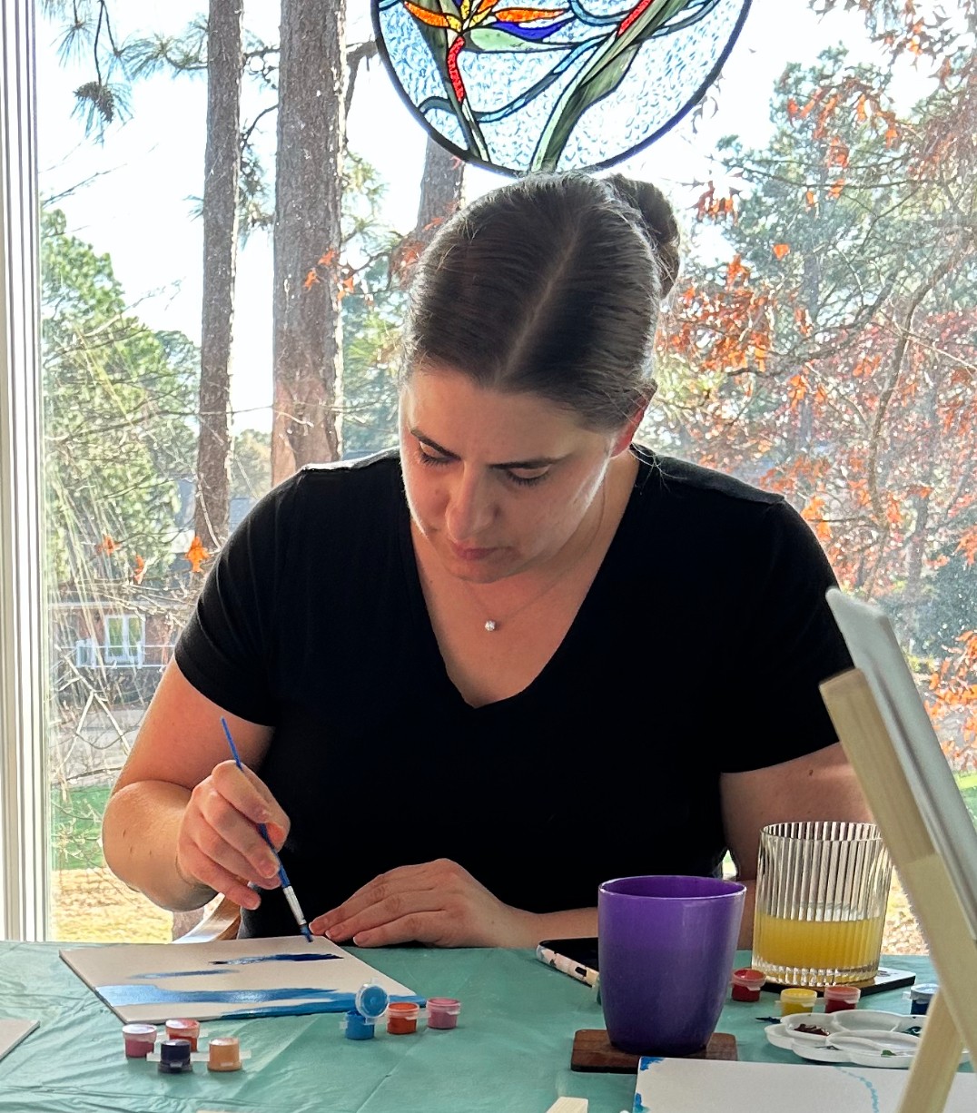 Woman with dark hair in a bun, wearing a black shirt, painting at a table with art supplies, window with trees outside