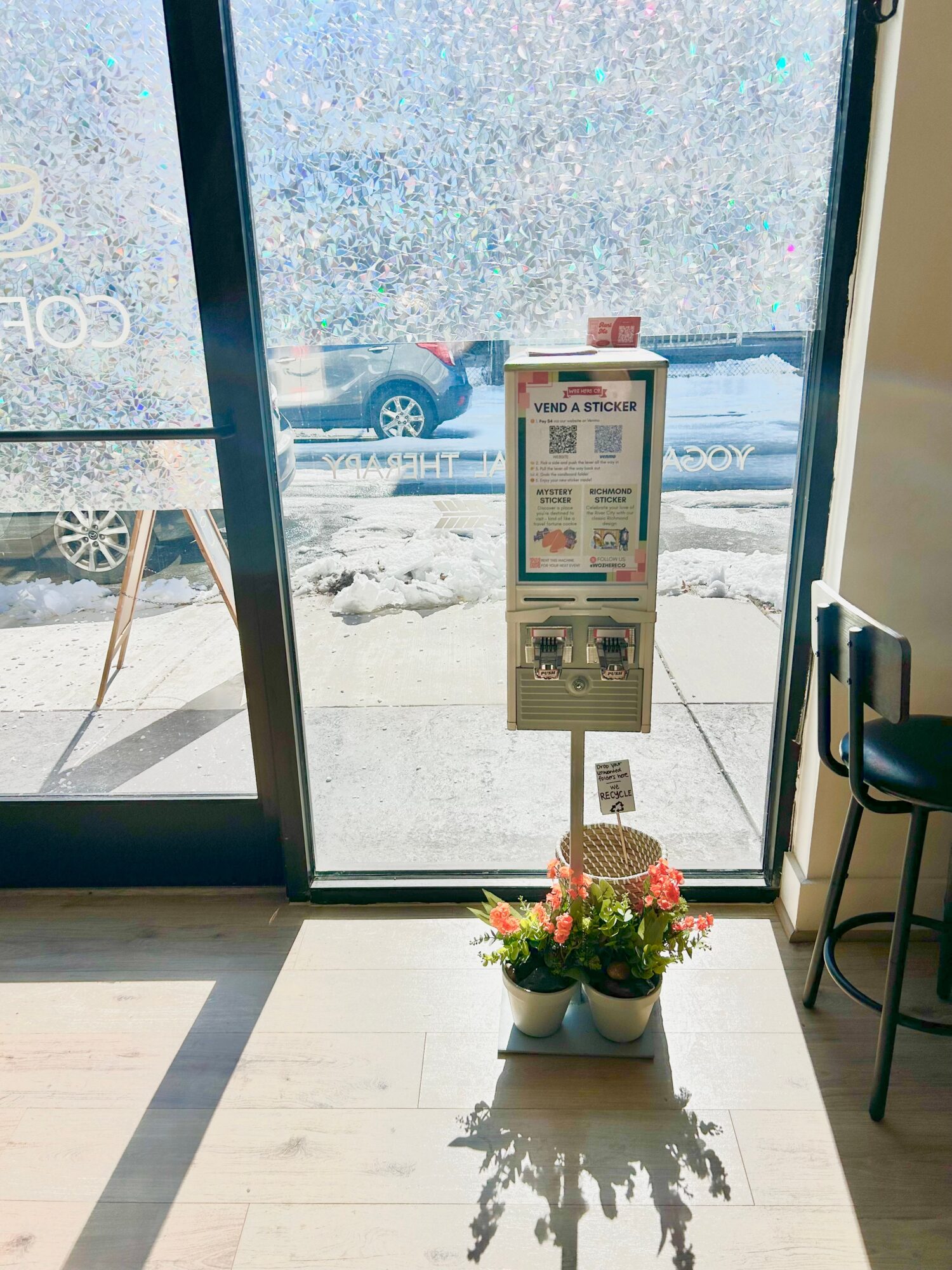 View through glass door showing snow outside, a vending machine inside, and a potted plant with flowers near the door.