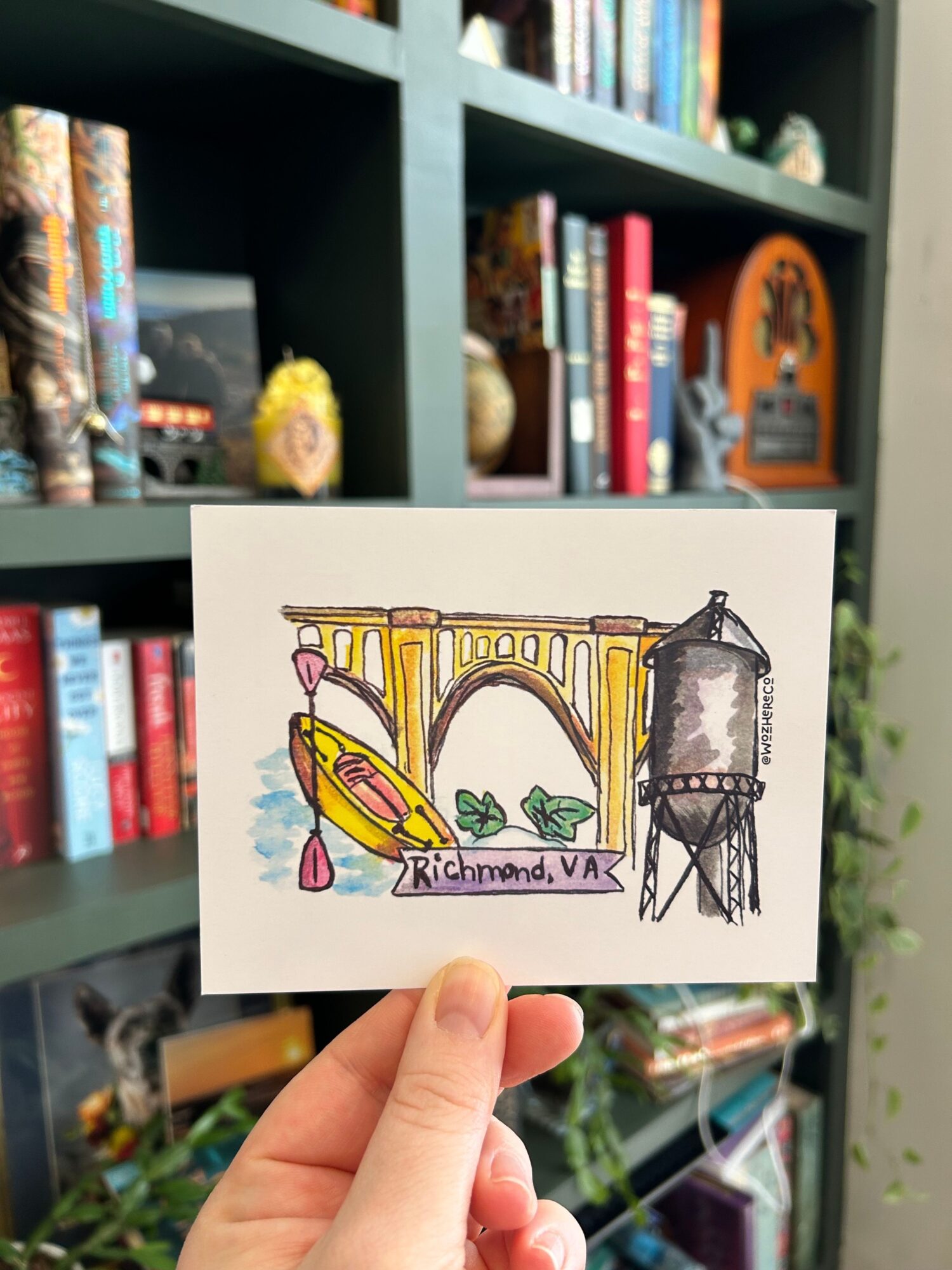 Hand holding a colorful drawing of a garden scene with a fountain, a bench, and a banana, in front of a bookshelf.