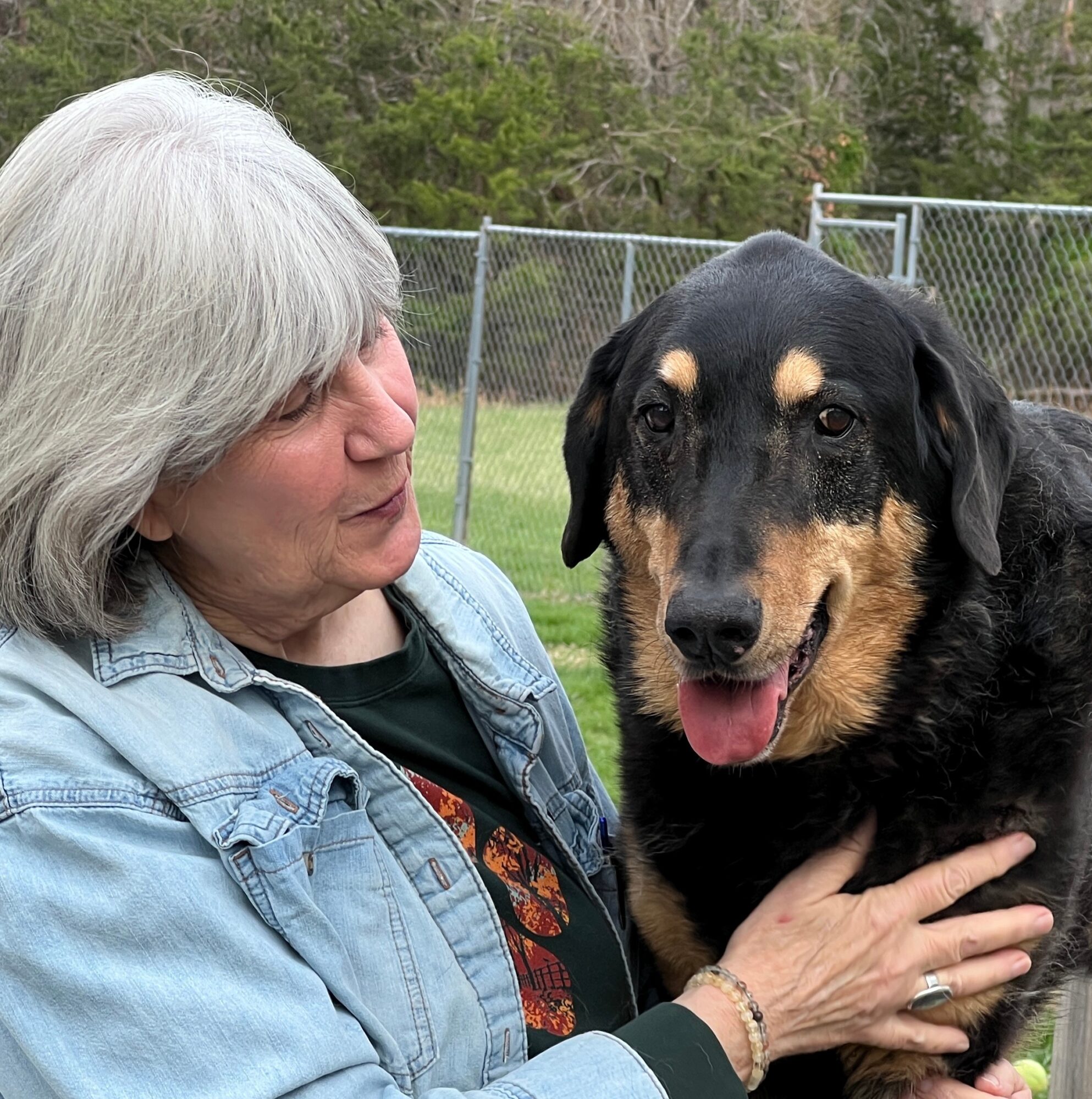 Woman with gray hair holding a black and tan dog outdoors near a fence, trees in background.