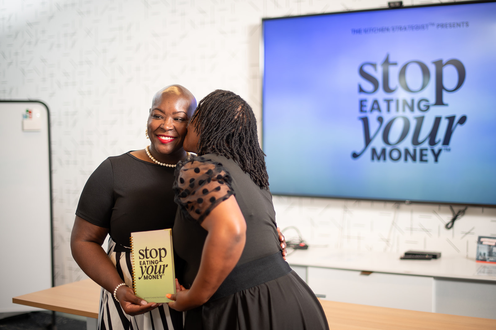 Two women smiling and talking, one holding a book titled 'Stop Eating Your Money,' in a room with a screen displaying a message.