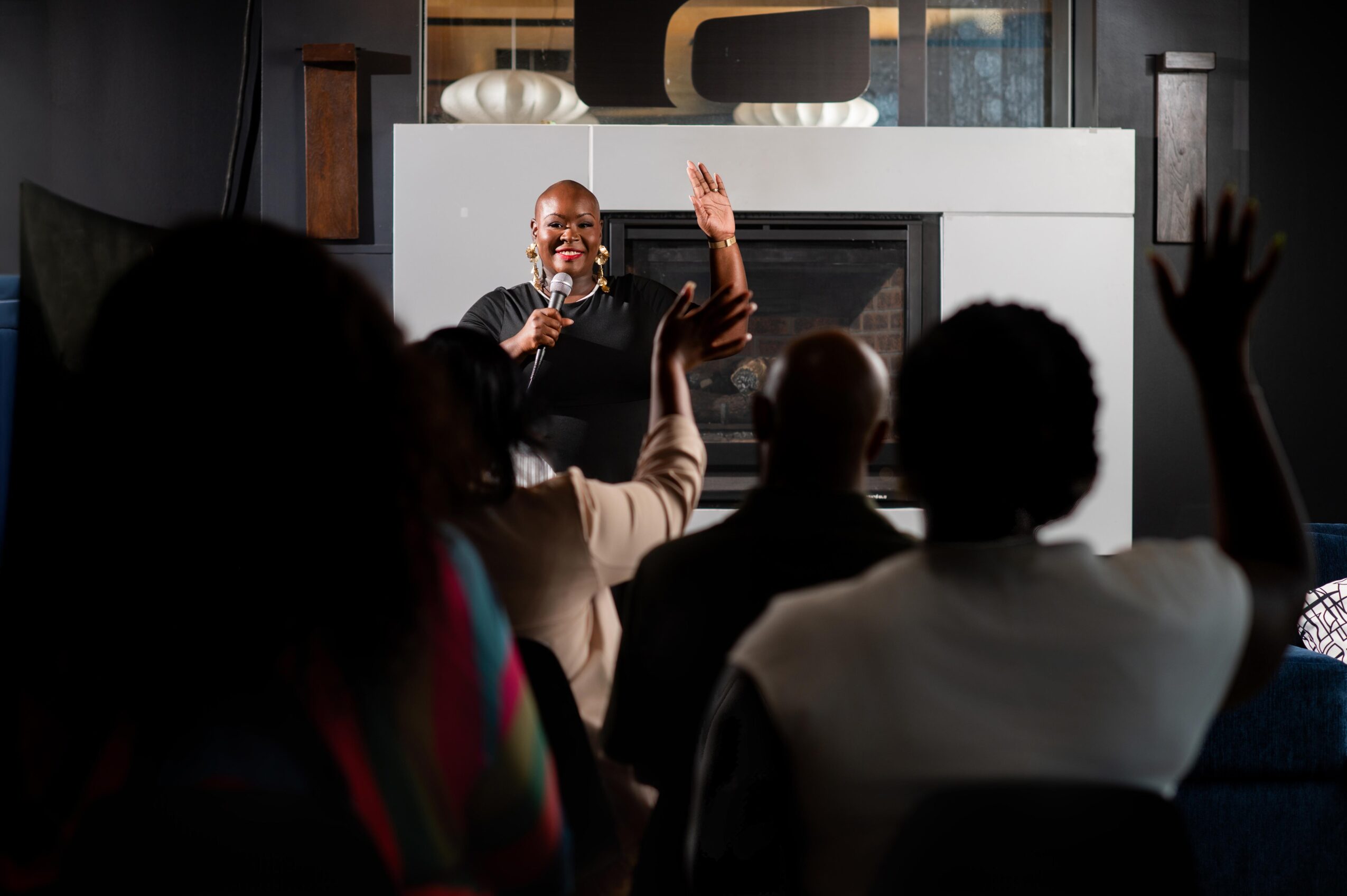 Person speaking on stage with microphone, audience members raising hands, in a dark room with a white background behind the speaker.