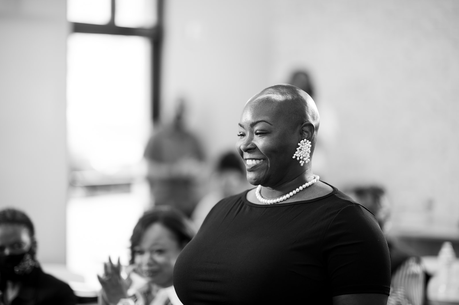 Smiling woman with earrings and necklace in a room with people, window in background.