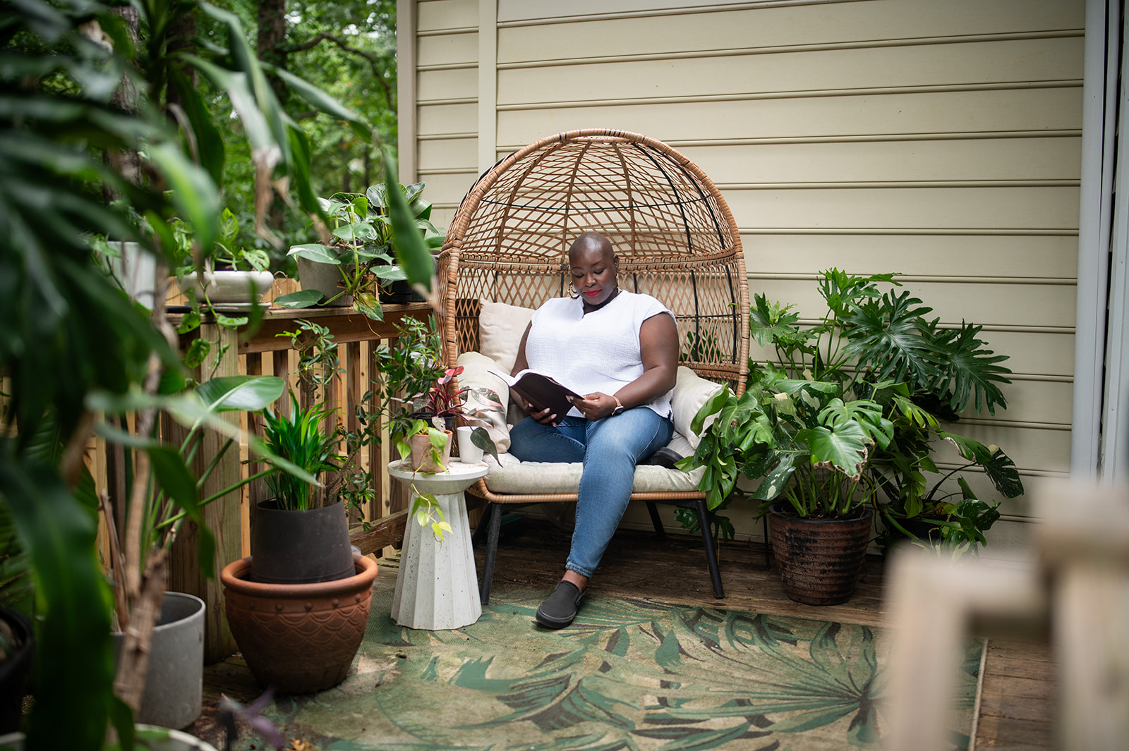 Person sitting on a wicker chair reading a book surrounded by potted plants on a porch.