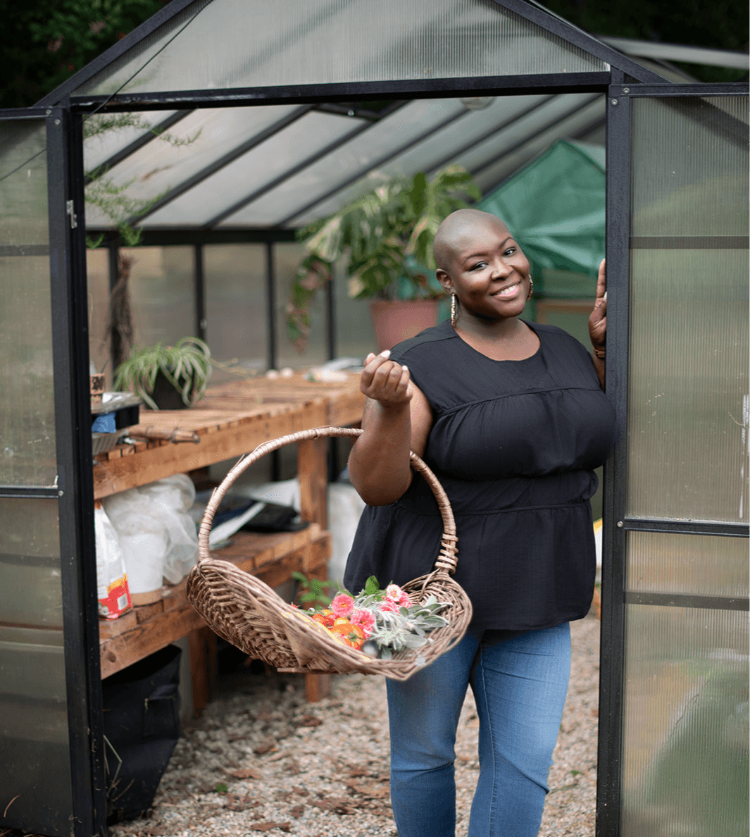Smiling woman holding a basket of flowers inside a greenhouse, with plants and gardening supplies visible behind her.