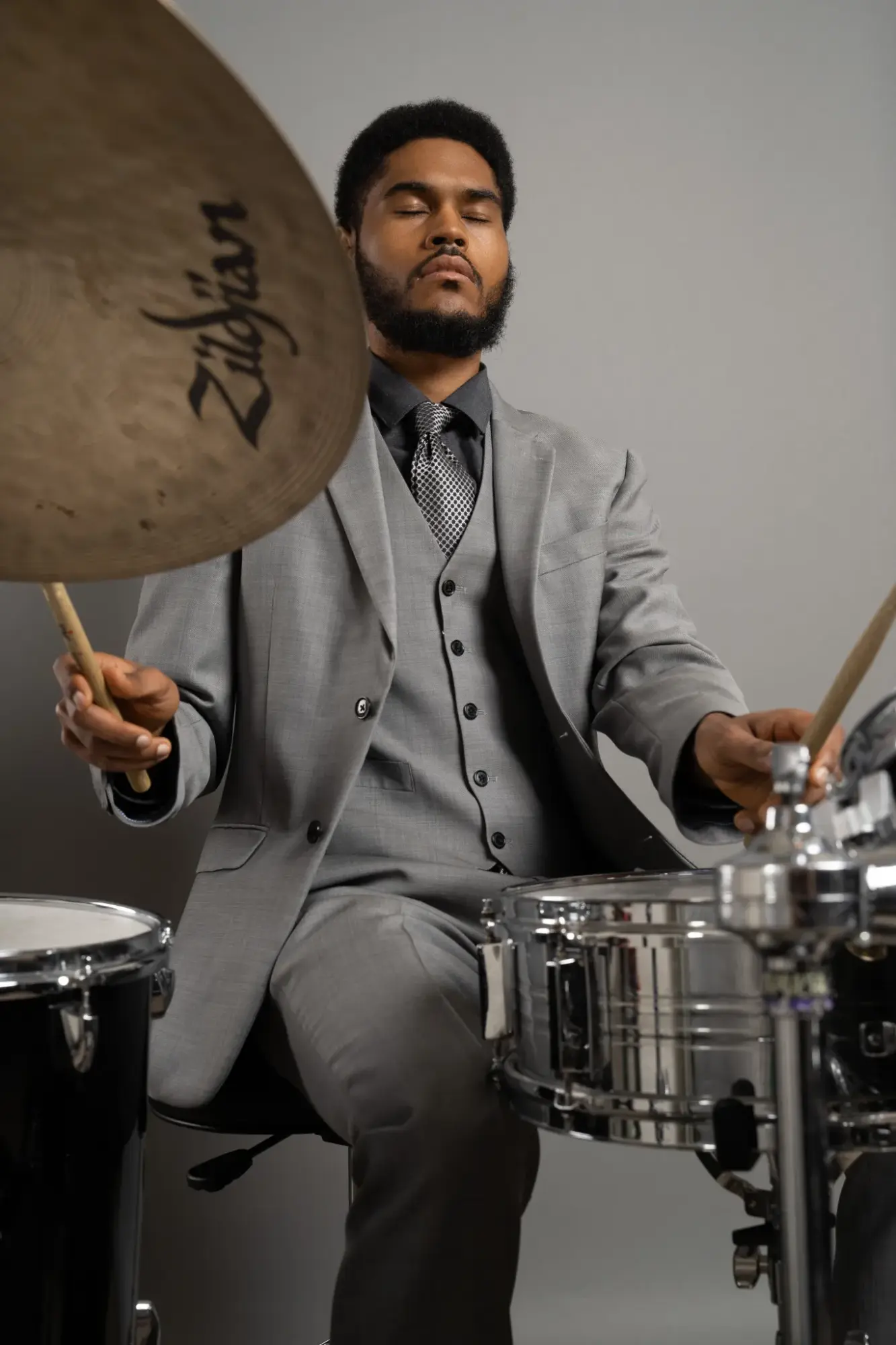 Man in gray suit playing drums, holding drumsticks, with a cymbal in the foreground, against a plain background.